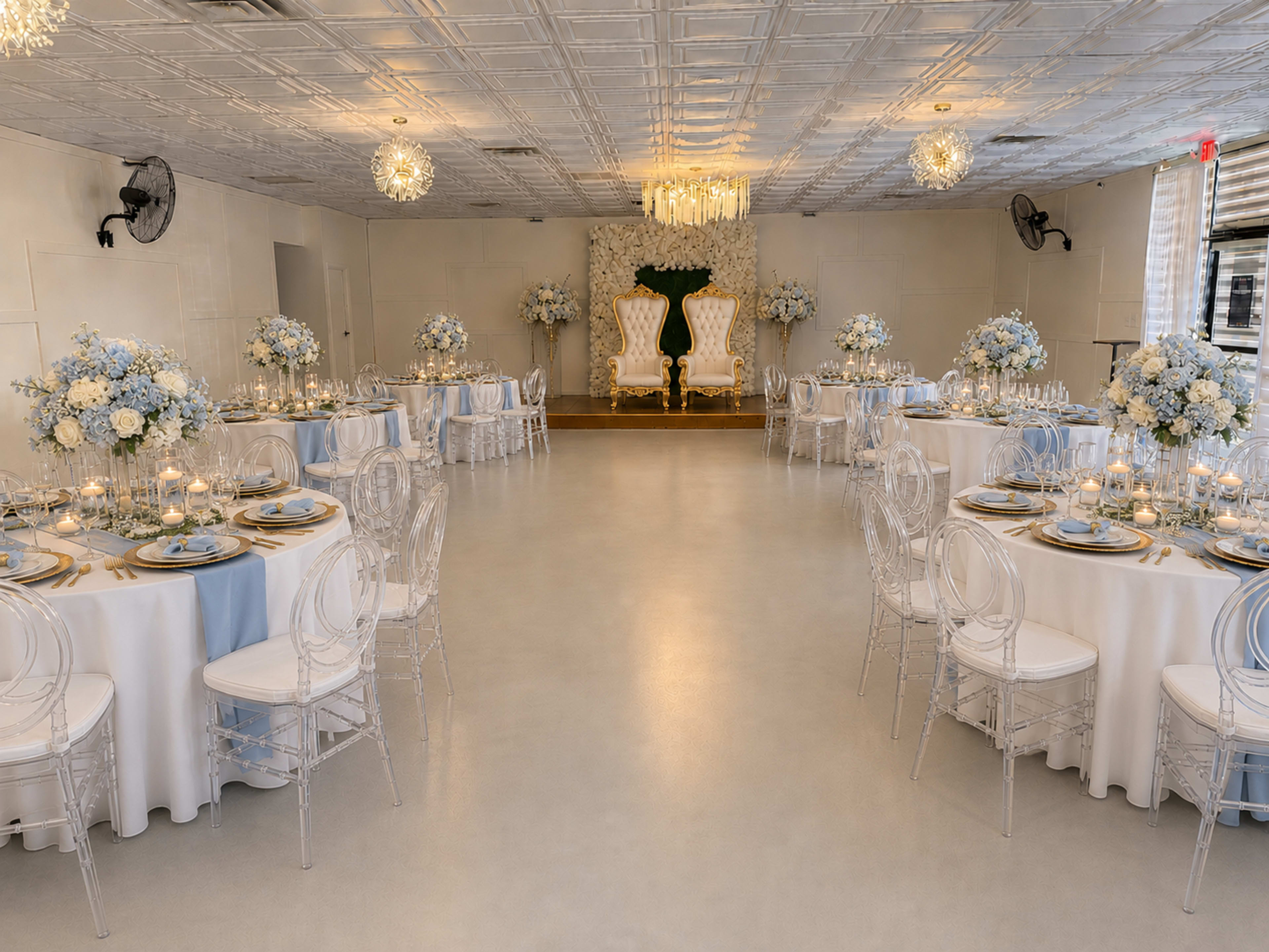 The image shows a banquet hall with elegantly set round tables adorned with floral centerpieces, while two ornate chairs are positioned at the front against a backdrop of greenery and white flowers.