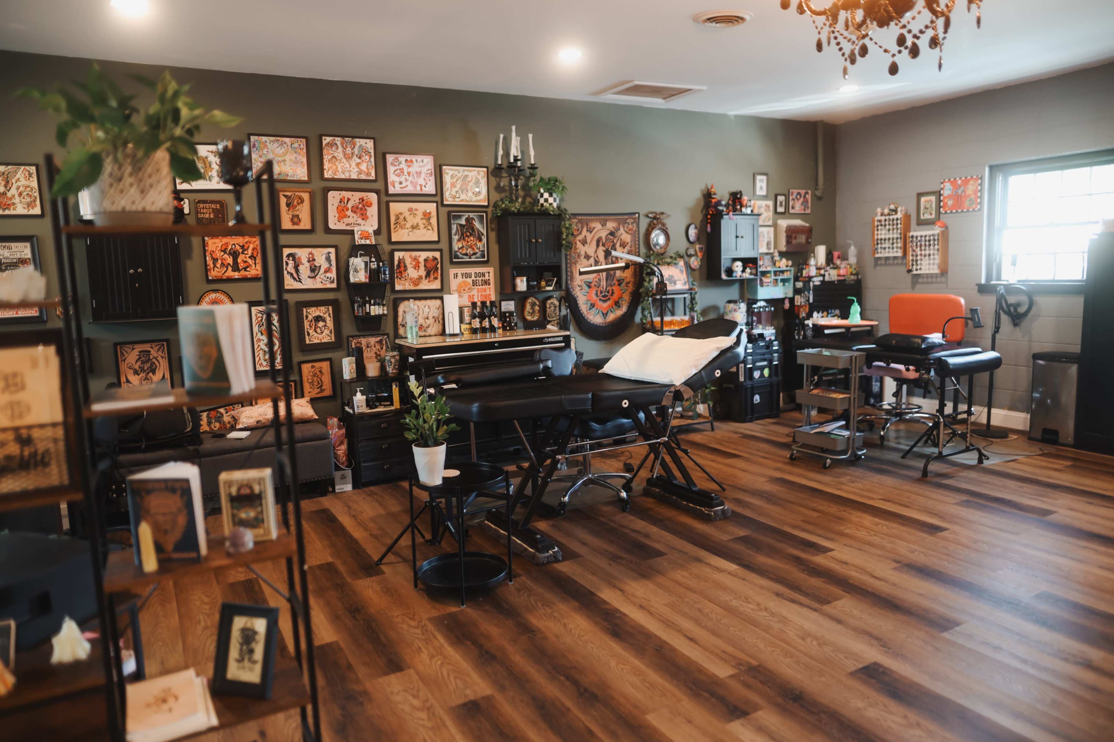 The image shows a tattoo studio featuring multiple workstations, decorated walls with framed artwork, and wooden flooring.