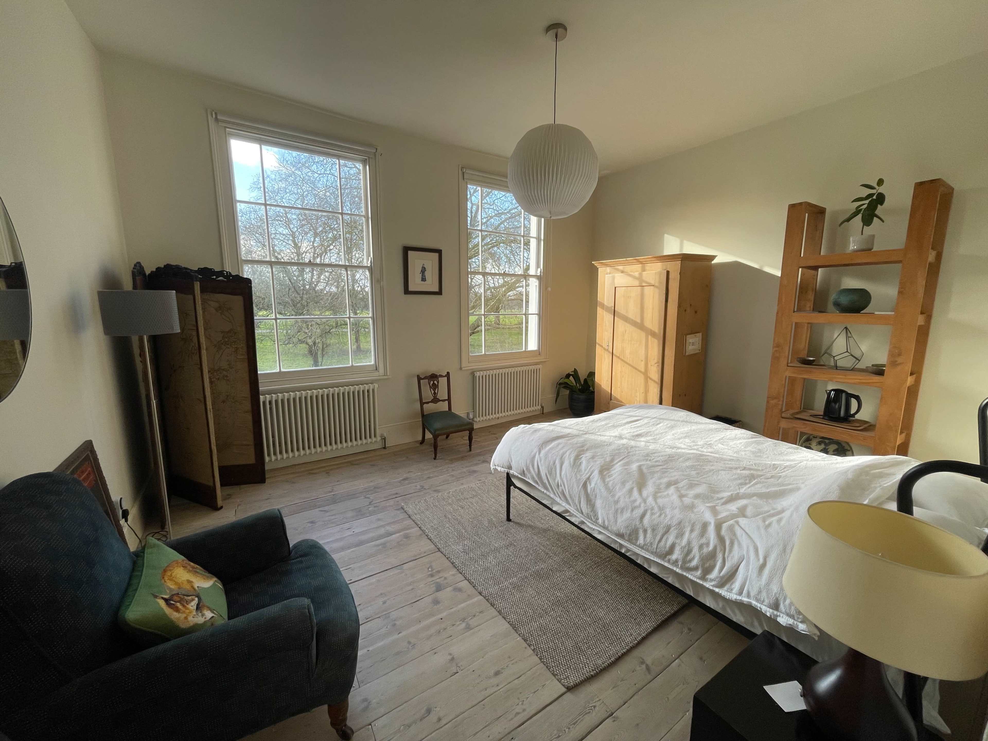 The room features a bed, a chair, a wooden cabinet, and large windows that offer a view of the outside.