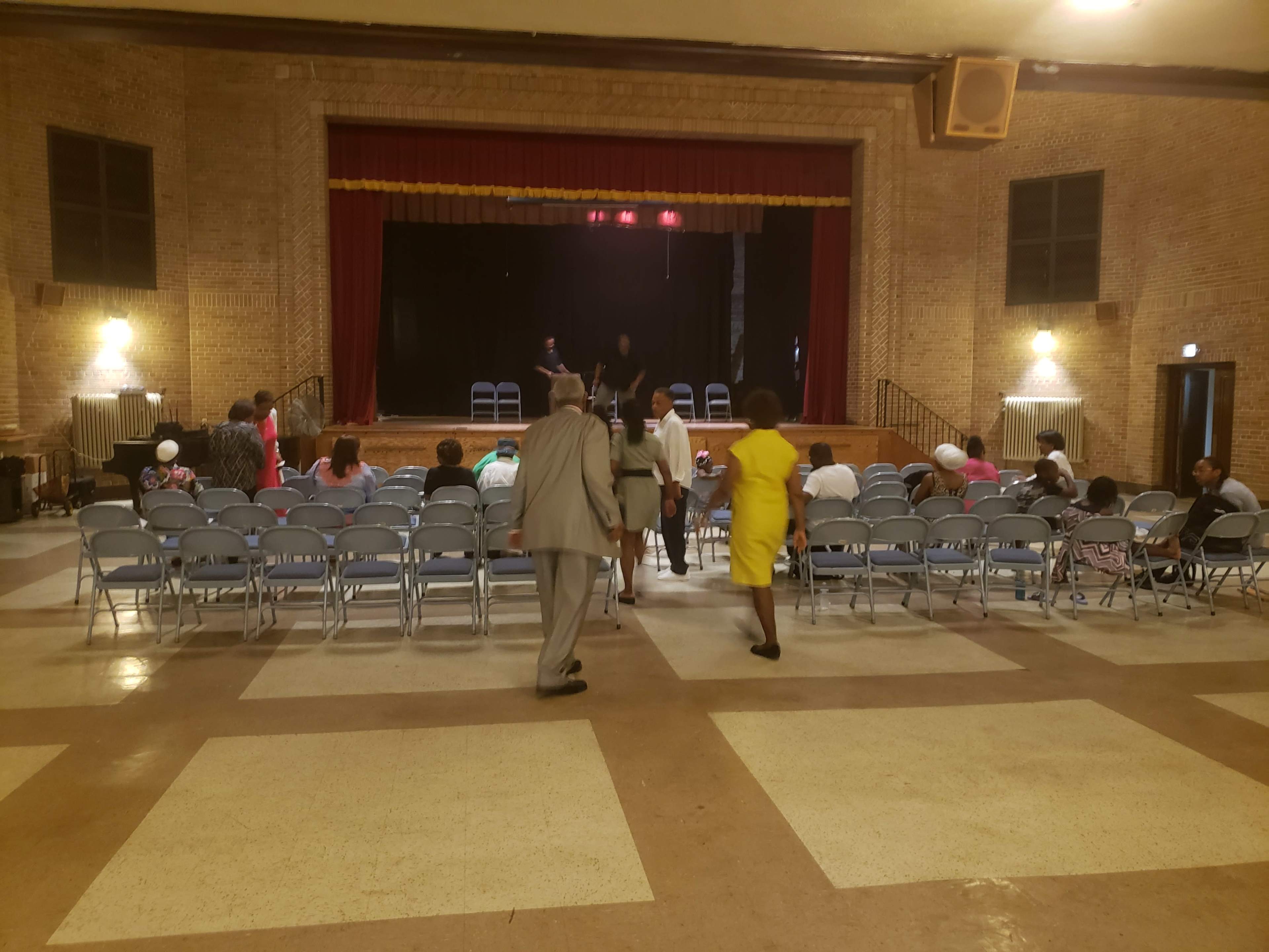 A group of people is entering a performance hall with rows of empty chairs facing a stage.