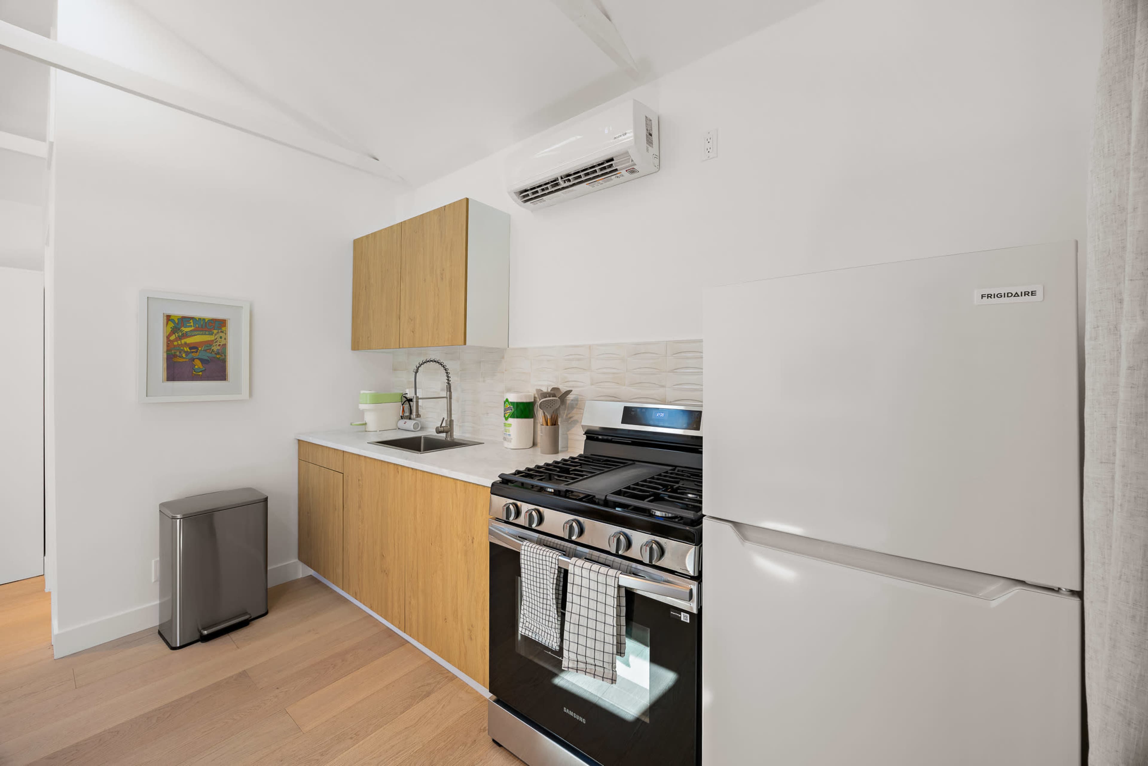 The image shows a modern kitchen with wooden cabinets, a stainless steel stove, a white refrigerator, and a small sink, all arranged in a compact space.