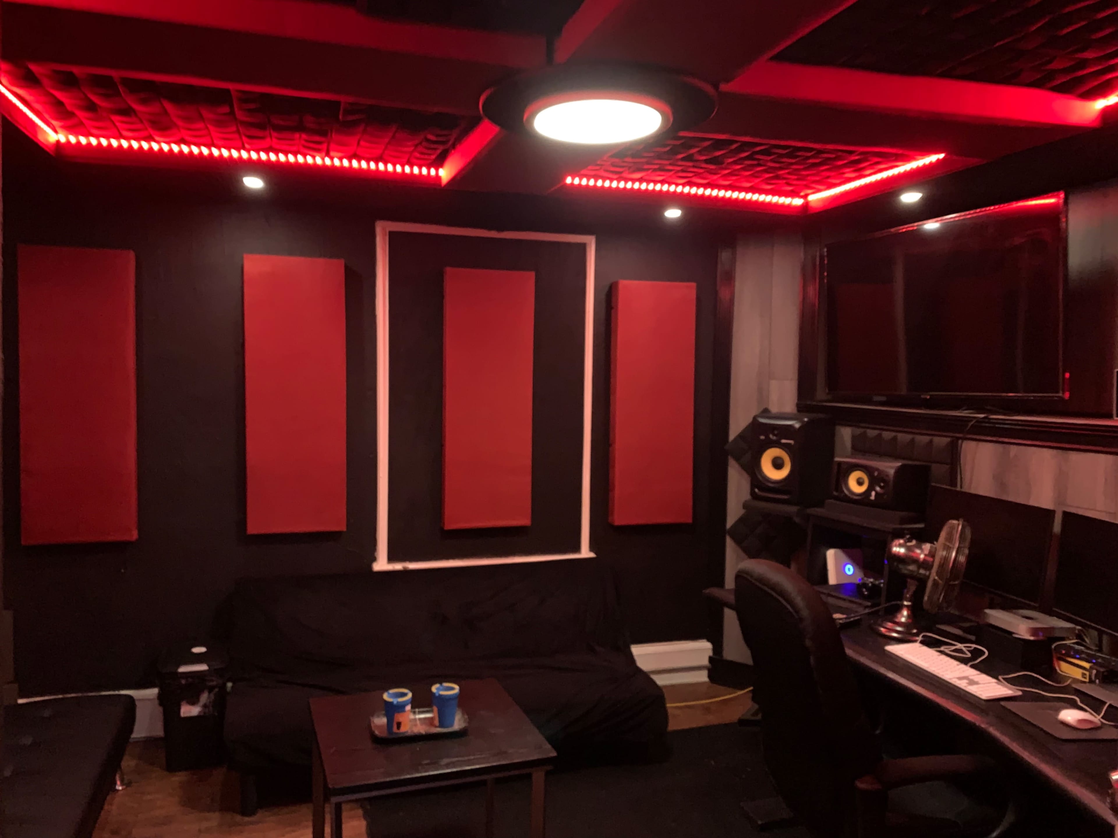 A dimly lit music studio with acoustic panels on the walls, a black sofa, and a desk with studio monitors and recording equipment.