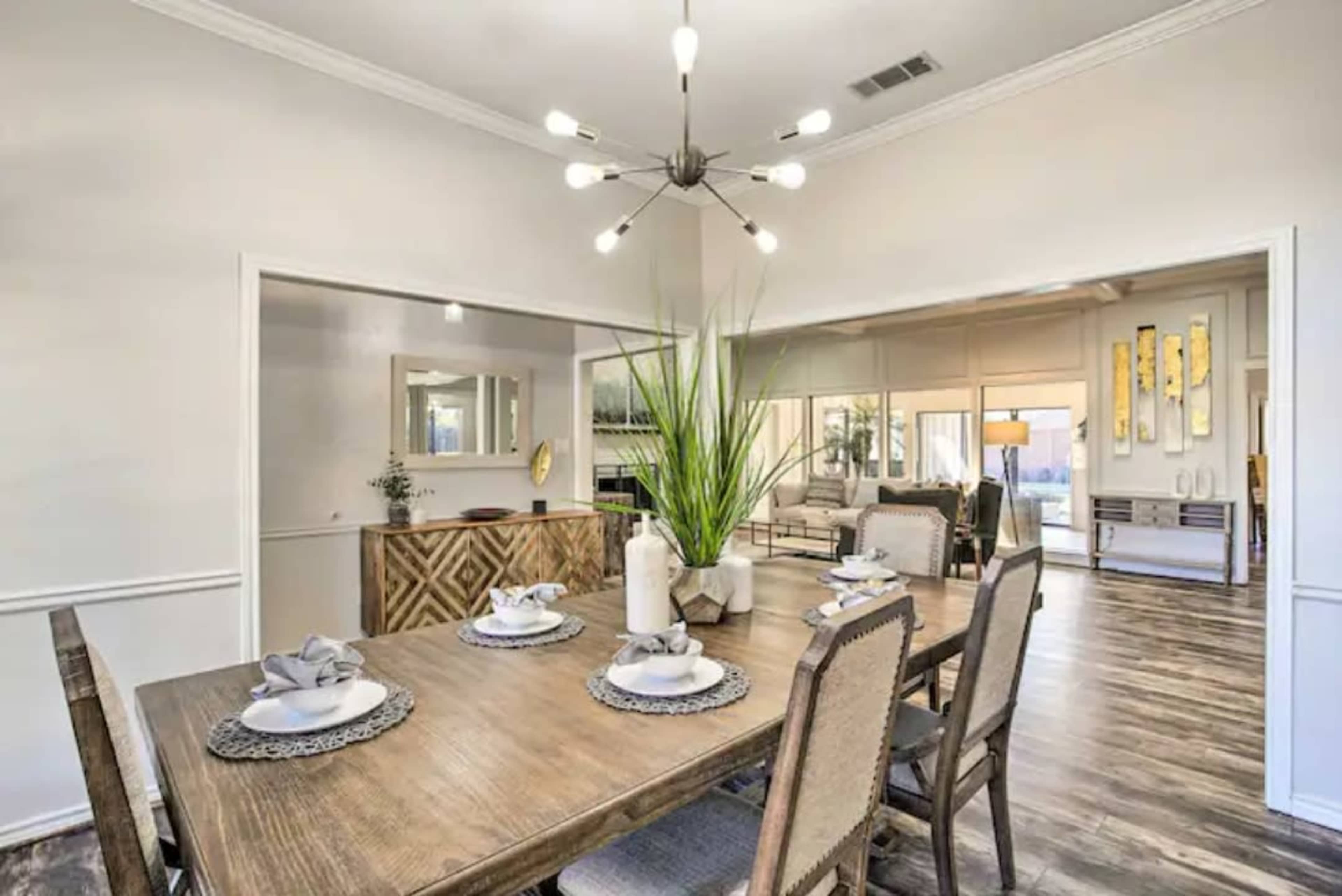 A dining area features a wooden table set with plates and a centerpiece, leading into a bright living space with large windows.