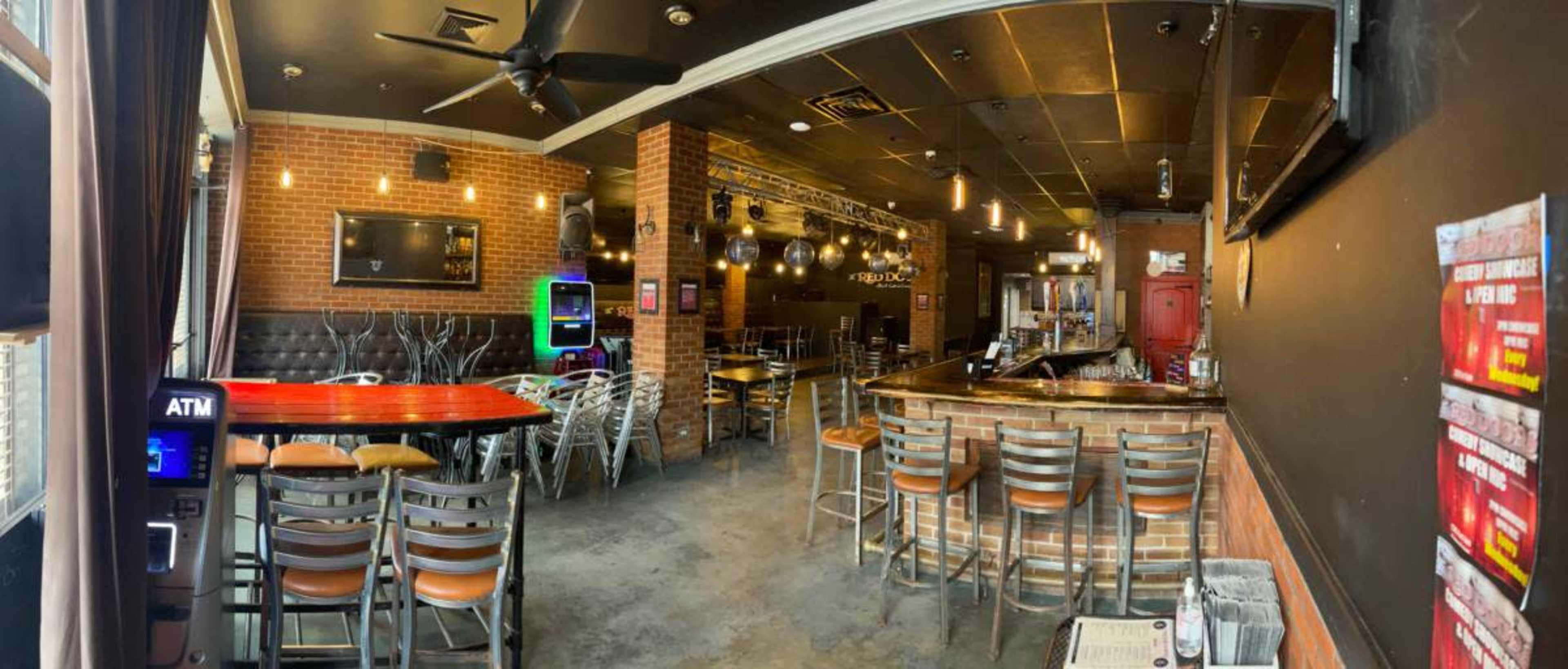 The image shows an interior of a restaurant or bar featuring a mix of seating arrangements, a bar area, and brick walls.