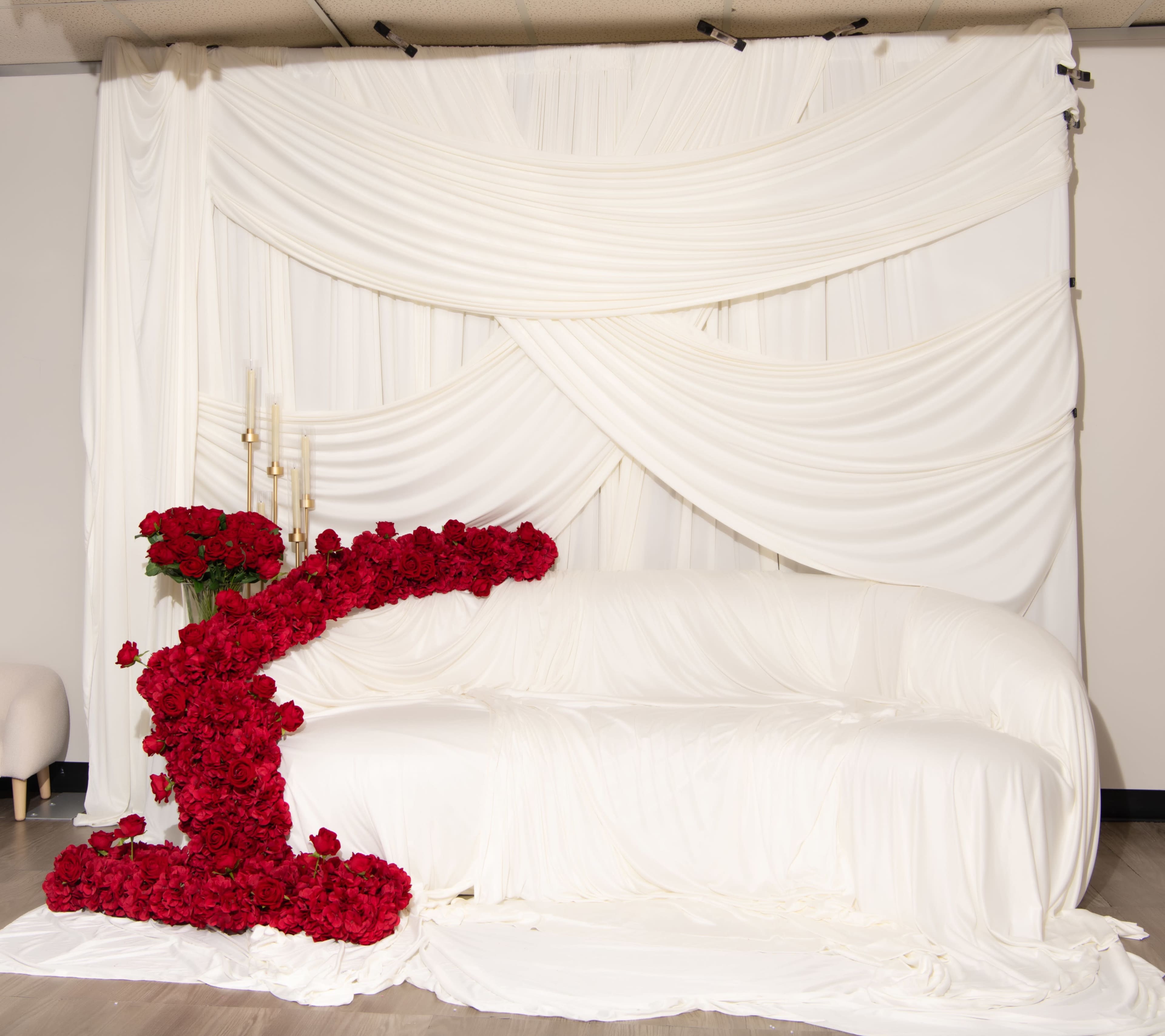 The image features a white sofa draped in a white fabric backdrop with a large arrangement of red roses shaped like the letter "C" positioned next to it.