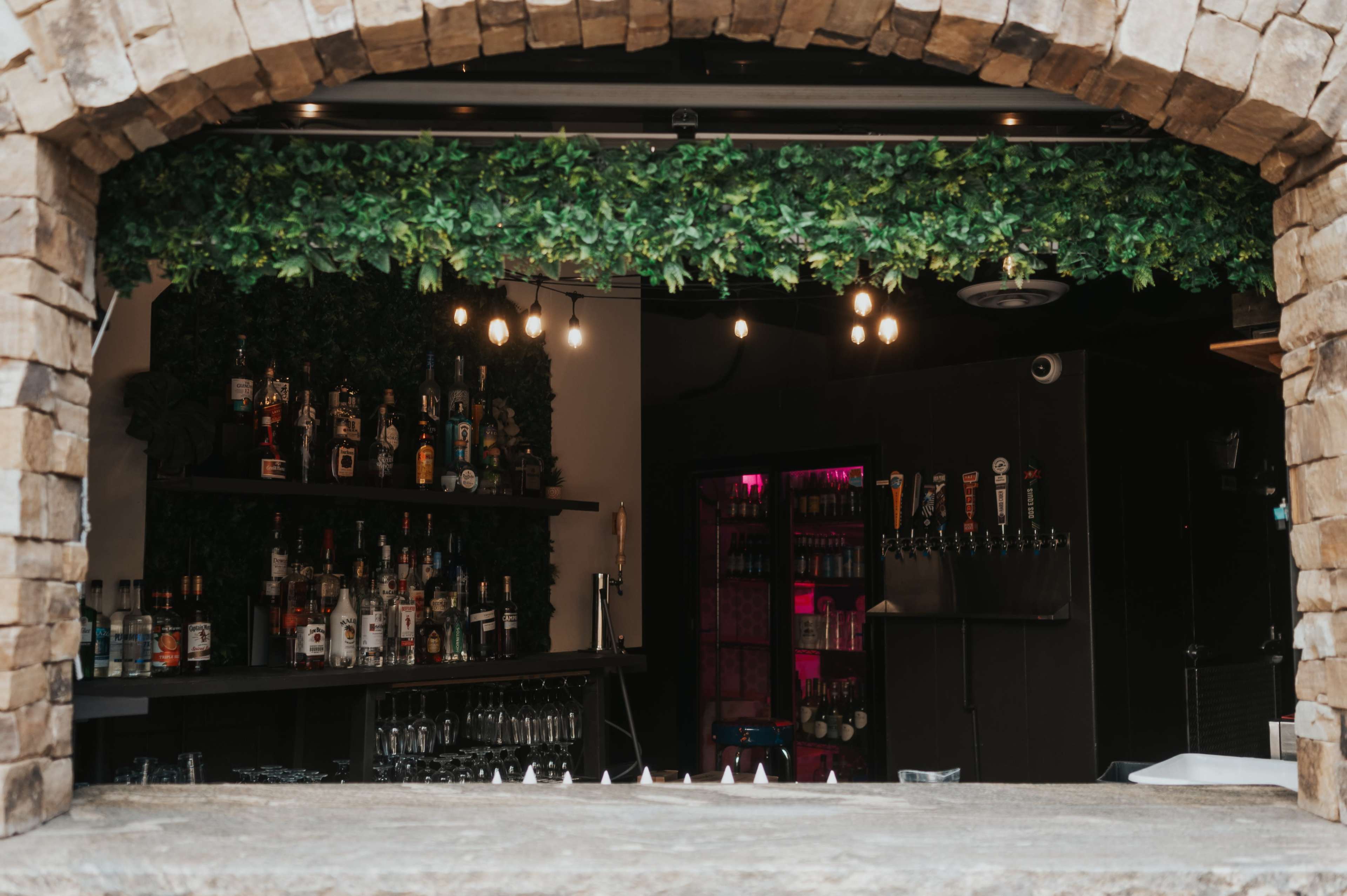 The image shows a bar with stone arch framing, featuring shelves stocked with various bottles and illuminated by pendant lights.