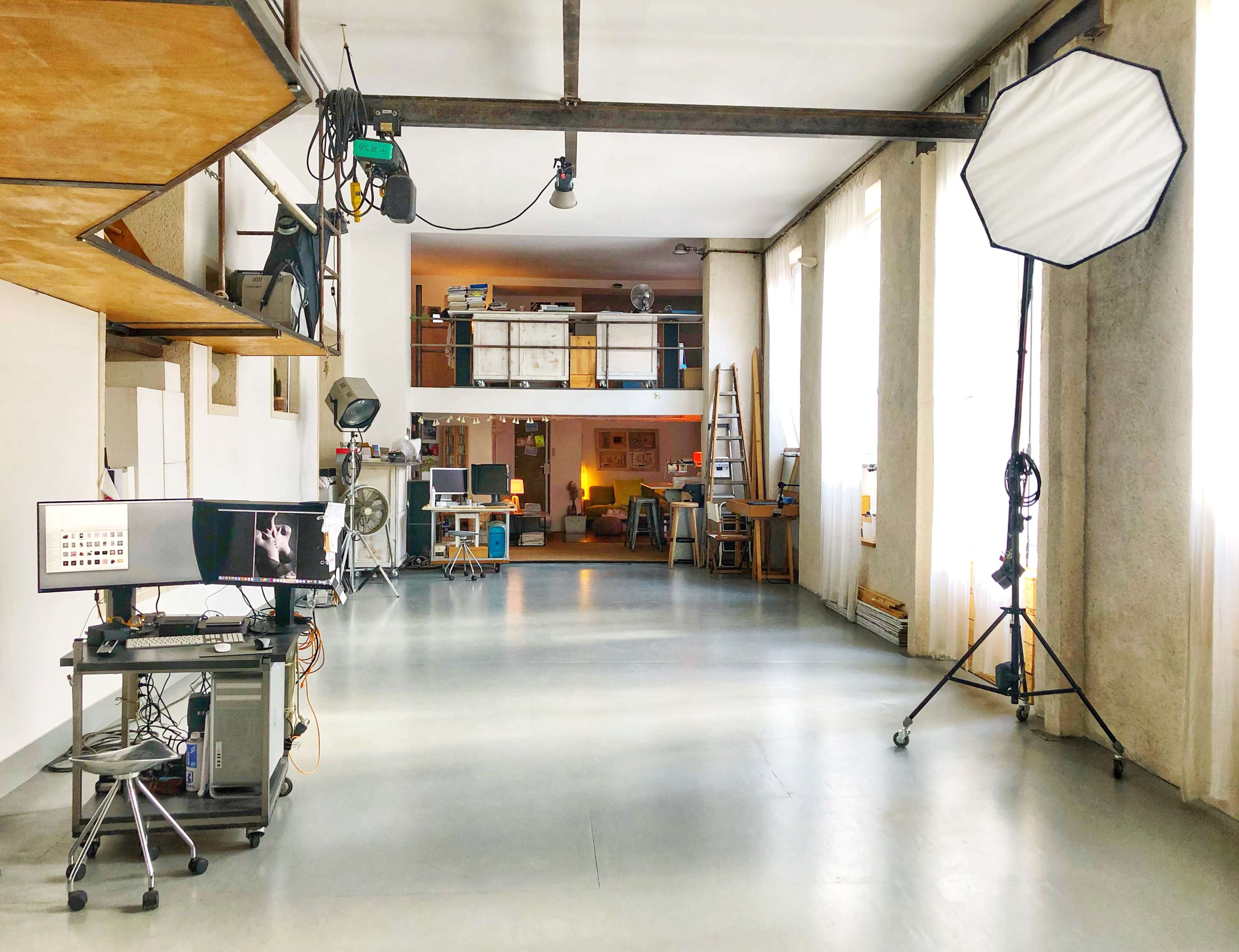 The image shows a spacious studio interior with large windows, equipment like lighting, a ladder, and various workspaces arranged in a minimalist layout.