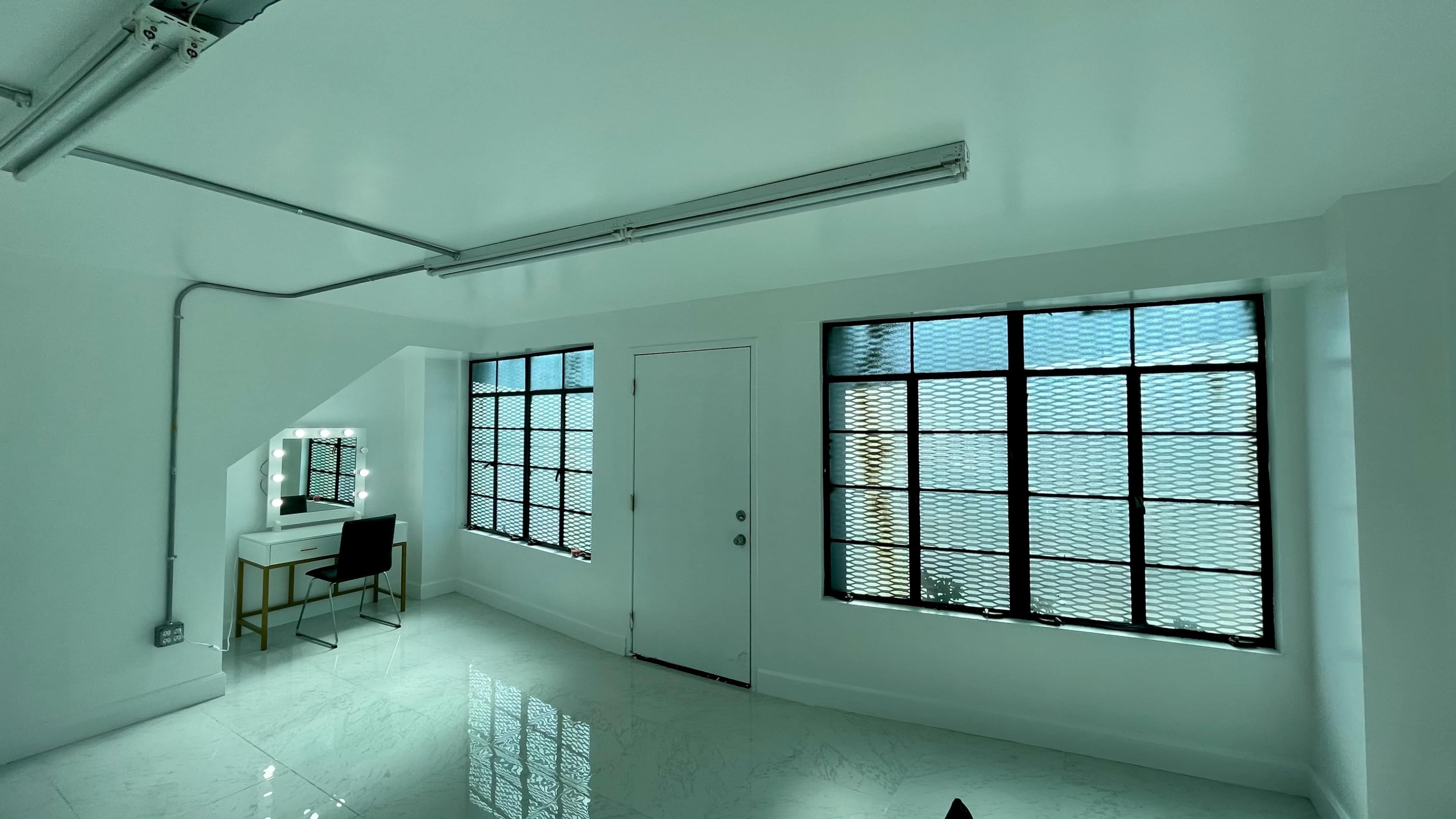 A bright room with large windows, a white door, and a vanity table with a mirror and a chair.