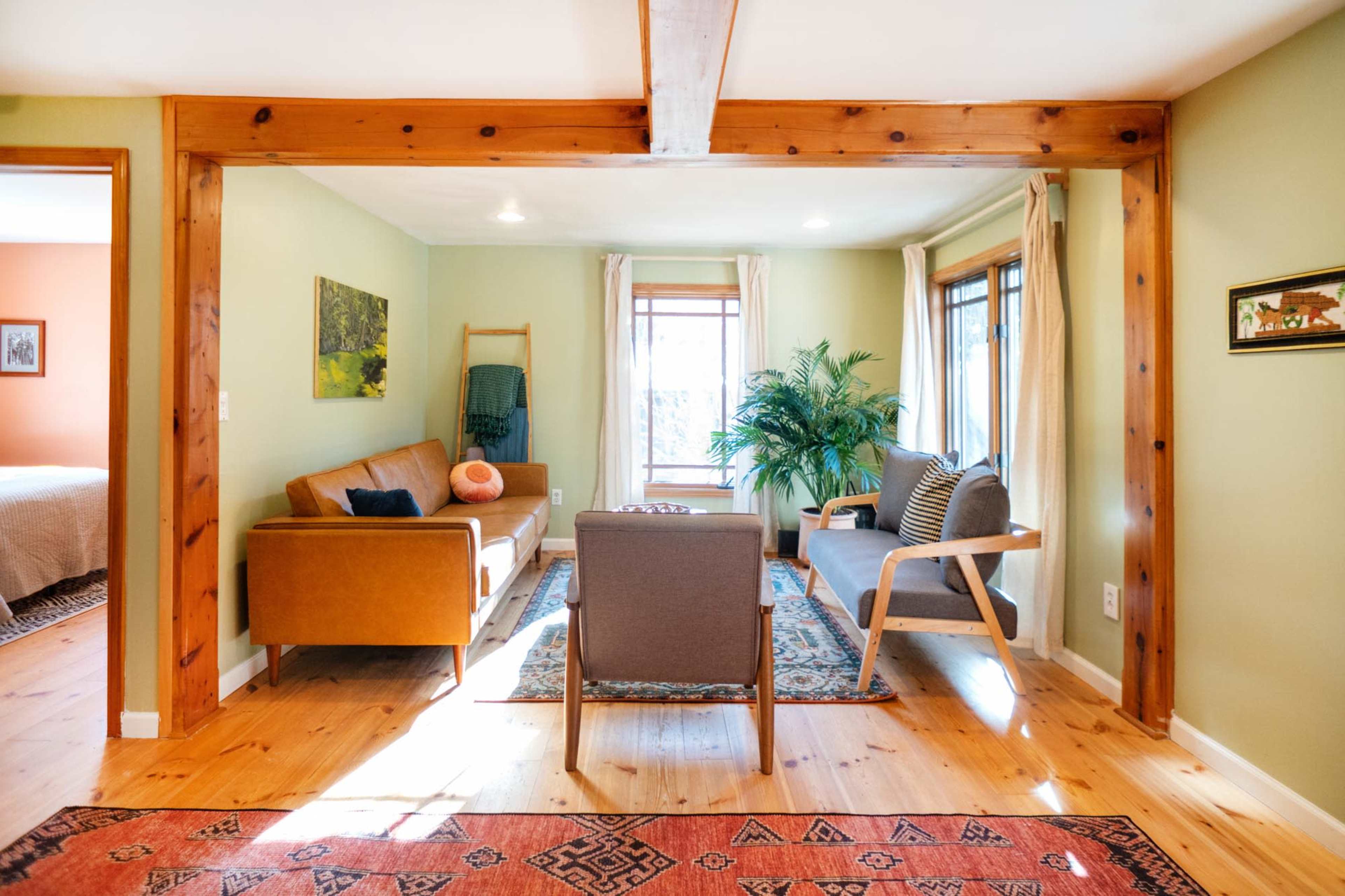 A cozy living room features a brown sofa, a pair of armchairs, a large window, and wooden beams, with light streaming in from outside.