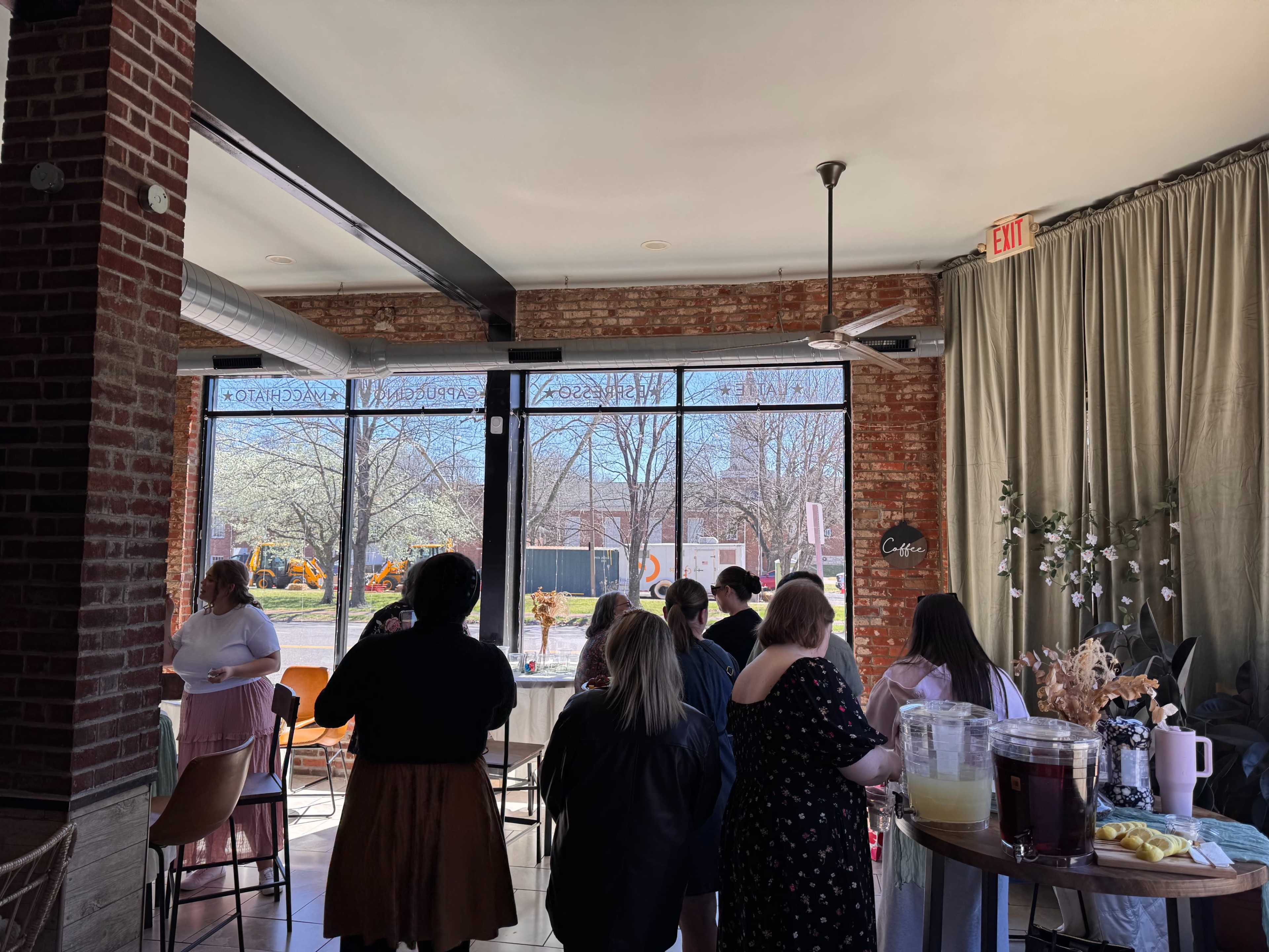 A group of people gathers inside a cafe featuring large windows that offer a view of a sunny outdoor scene.