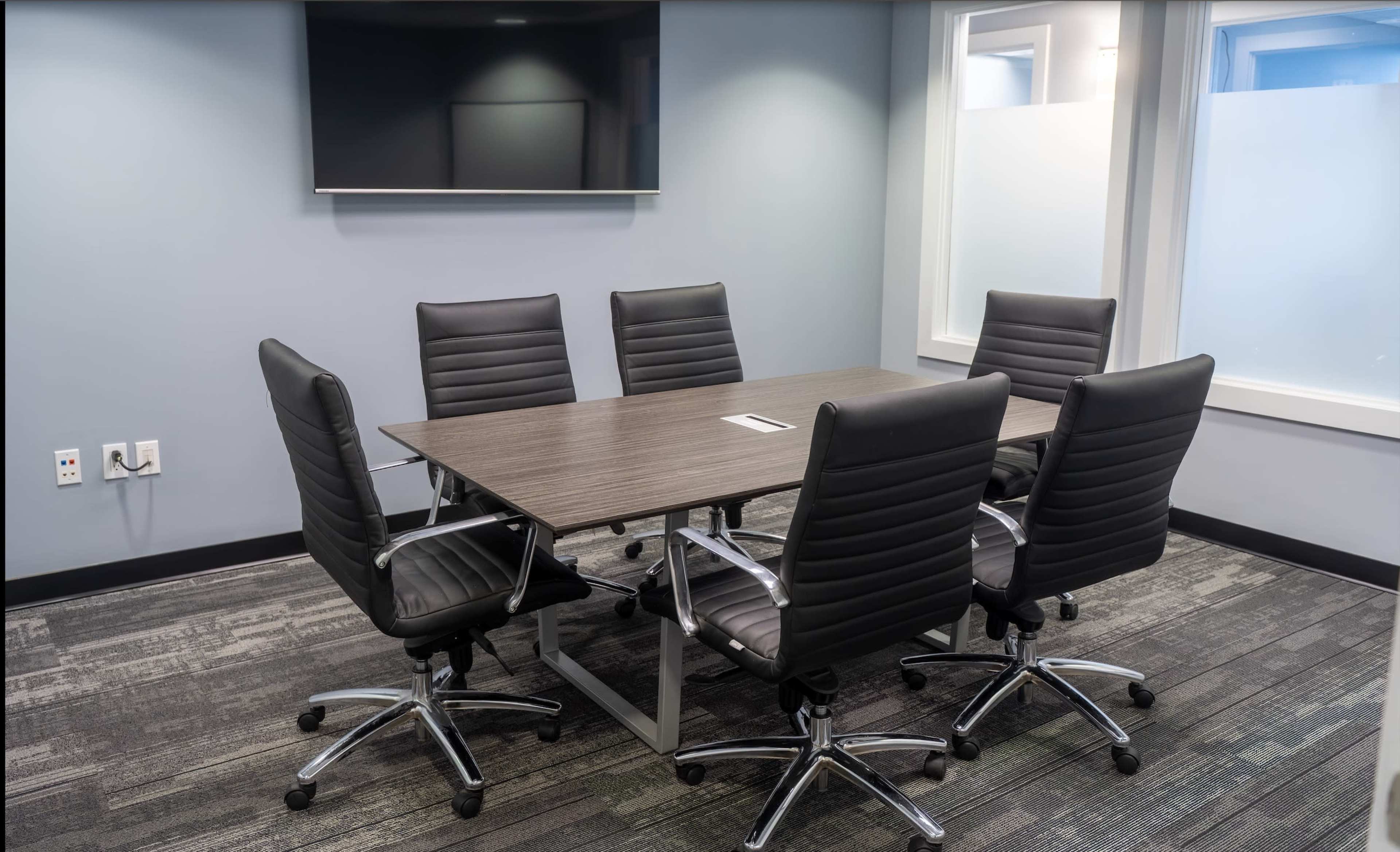 The image shows a modern conference room with a rectangular table surrounded by six ergonomic chairs and a television mounted on the wall.