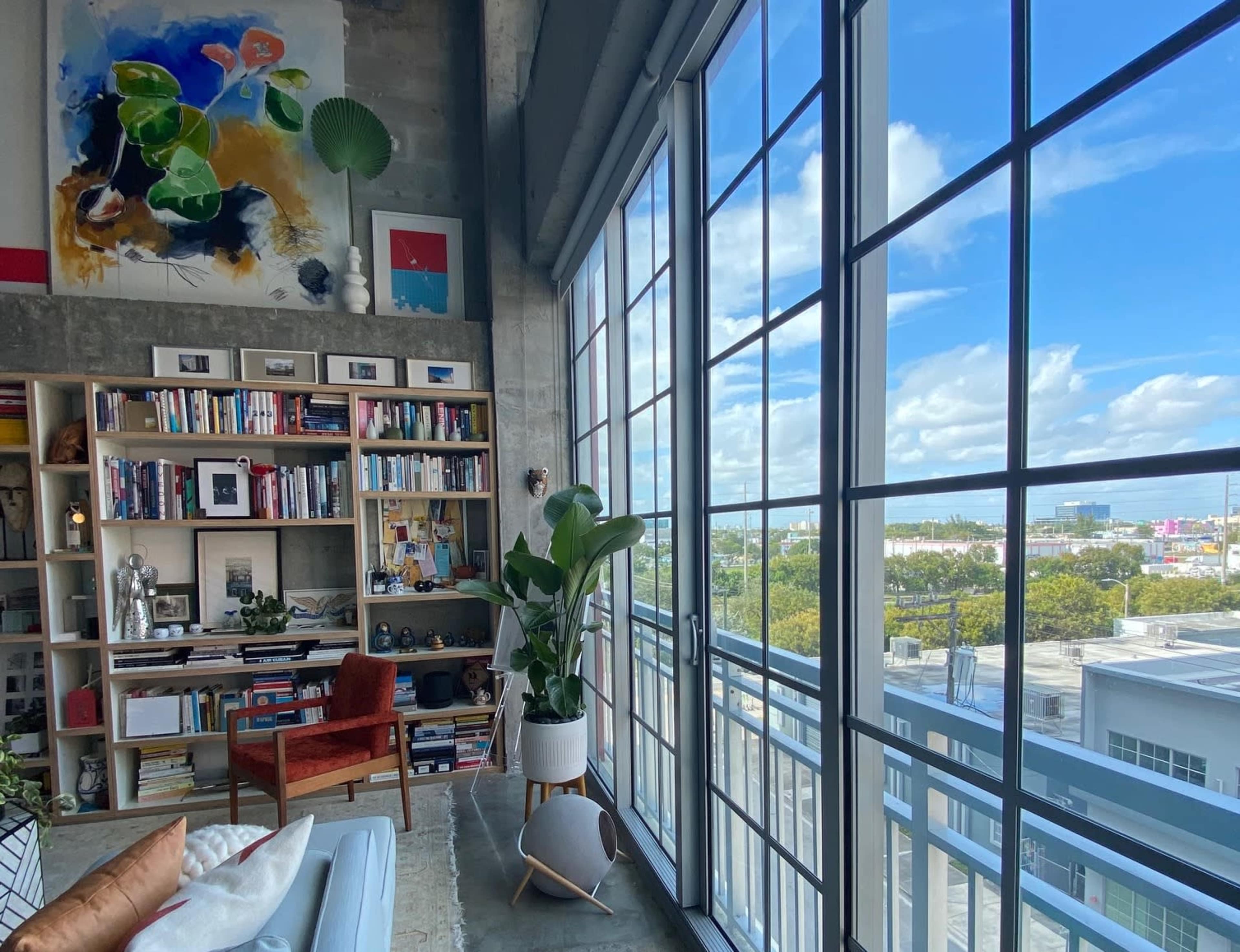 A modern living room features a large bookshelf, a comfortable armchair, and expansive windows with a view of the city skyline and blue sky.