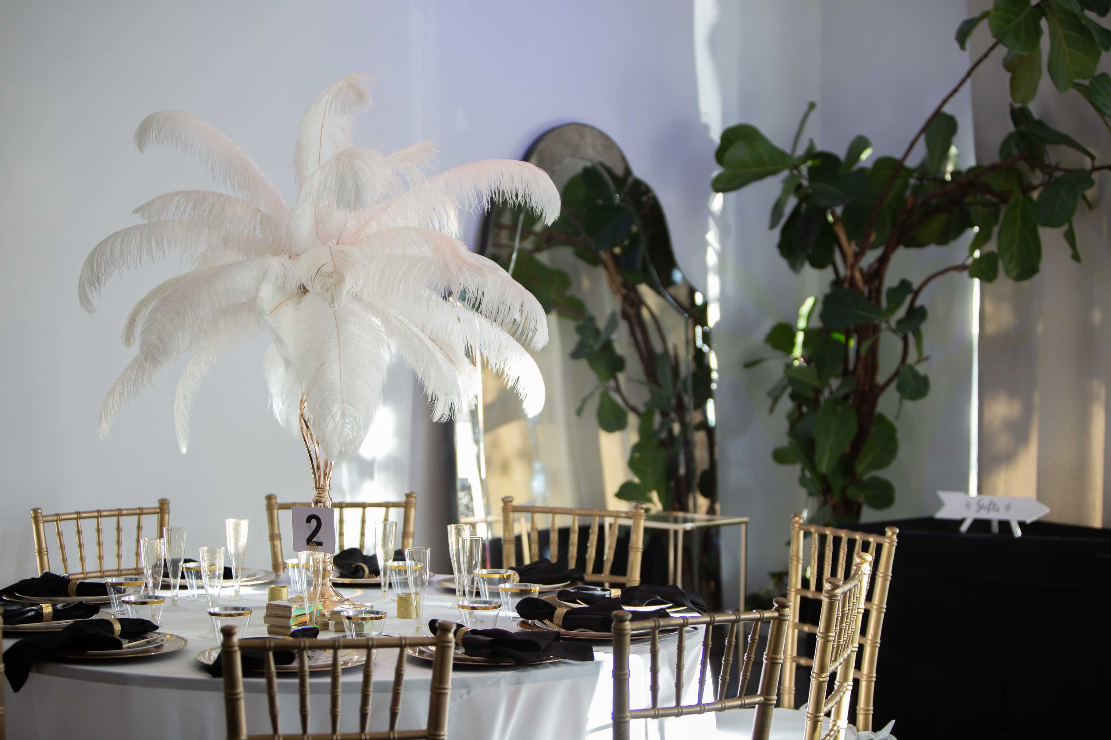 A elegantly set dining table features a large feather centerpiece, surrounded by gold chairs and illuminated by natural light.