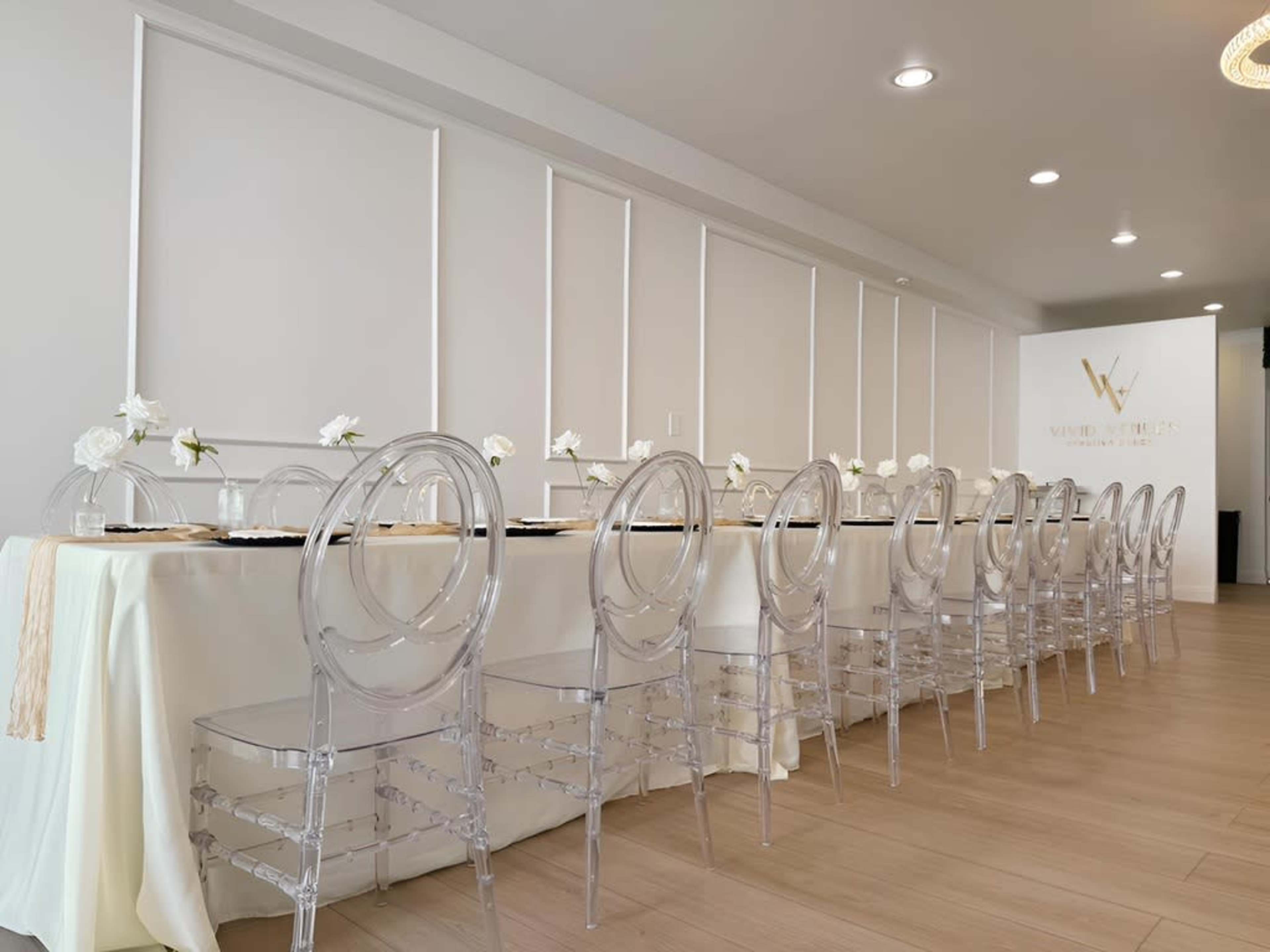 A long dining table is set against a neutral wall, adorned with clear chairs and white flowers as centerpieces.