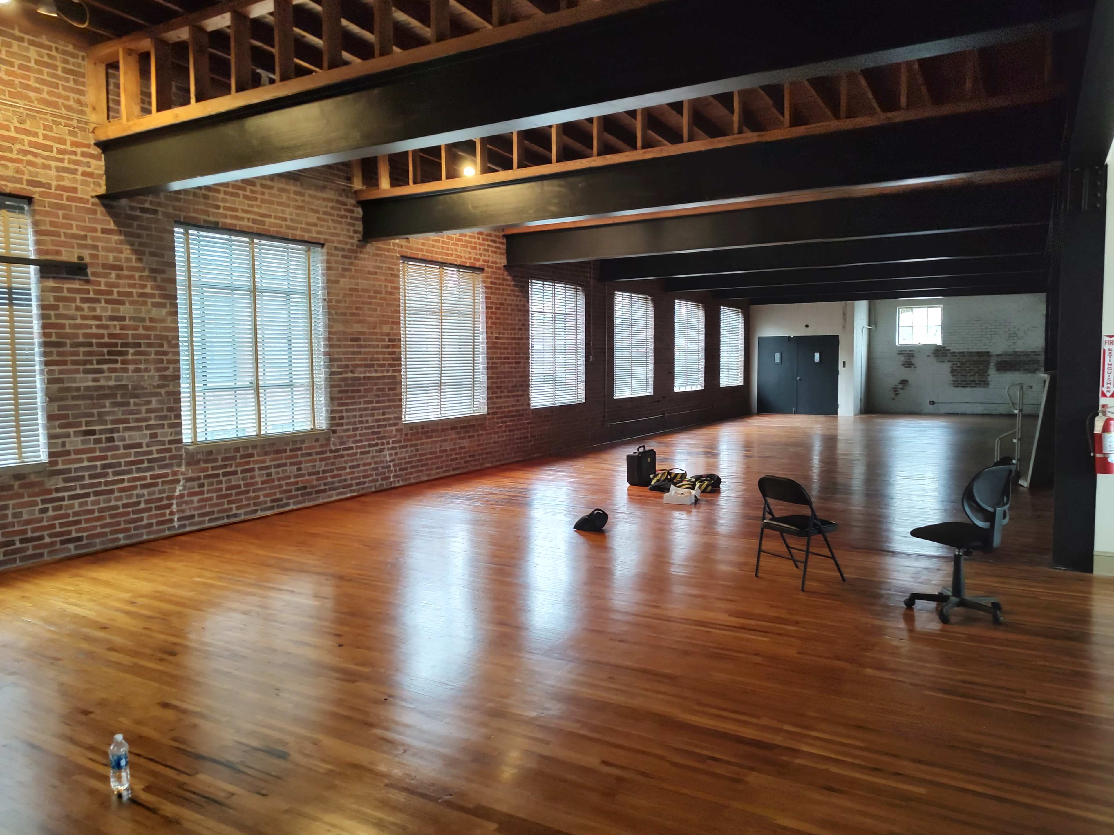 An empty industrial-style room with exposed brick walls, wooden floors, and large windows, featuring a few scattered chairs and personal items.