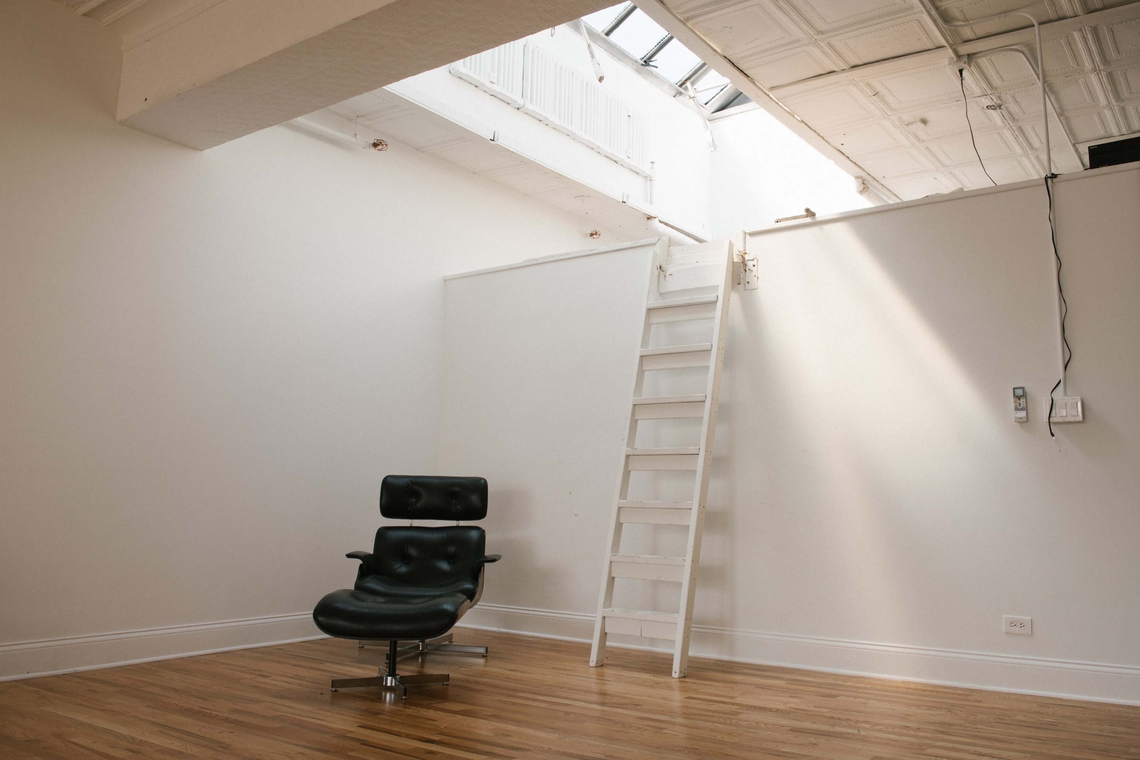 A minimalist room with a black leather chair and a white wooden ladder leading to a loft area.