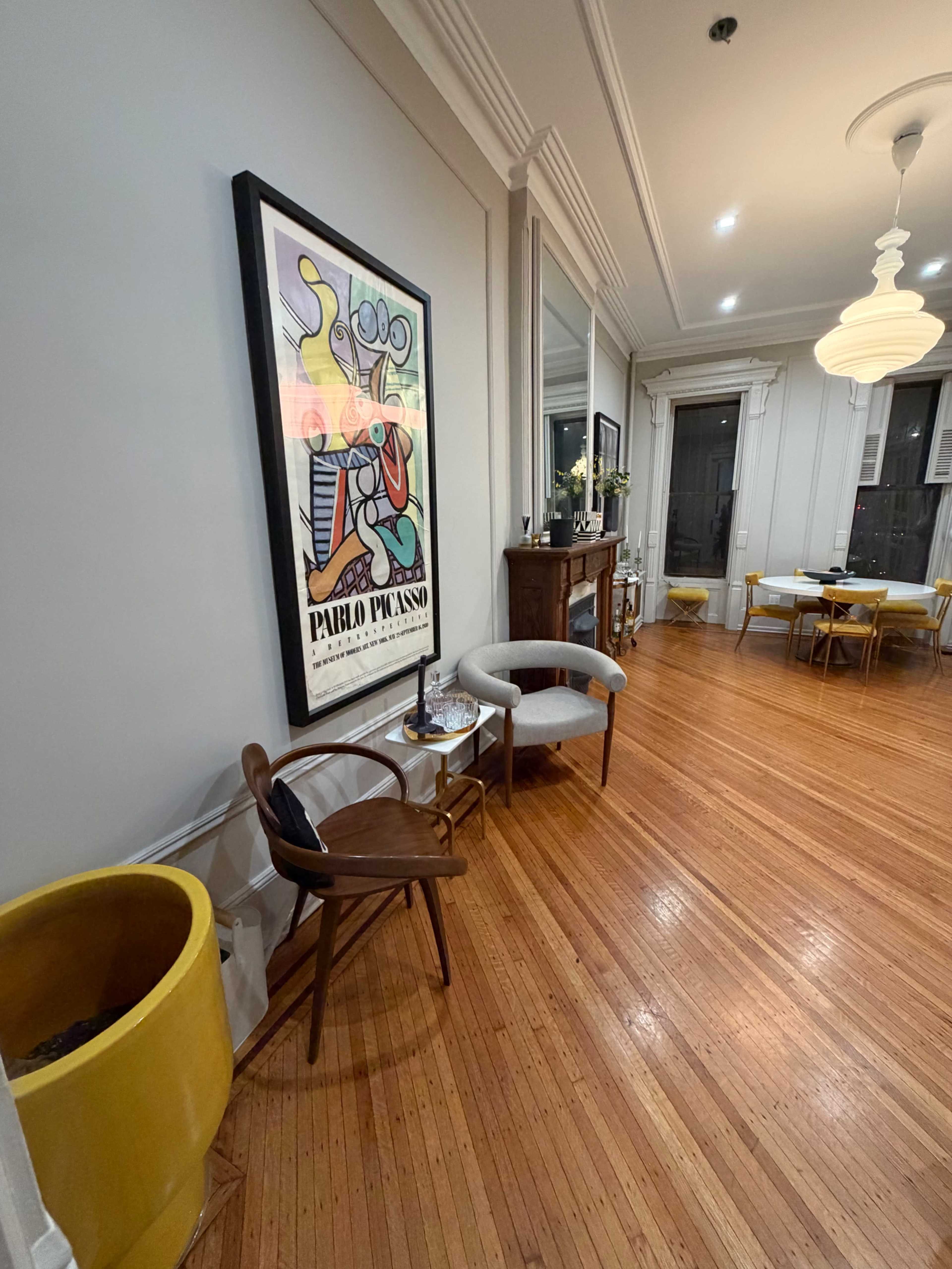 The image shows a room with polished hardwood floors, featuring a large framed poster on the wall, a yellow planter, a wooden chair, and a table set for dining in the background.