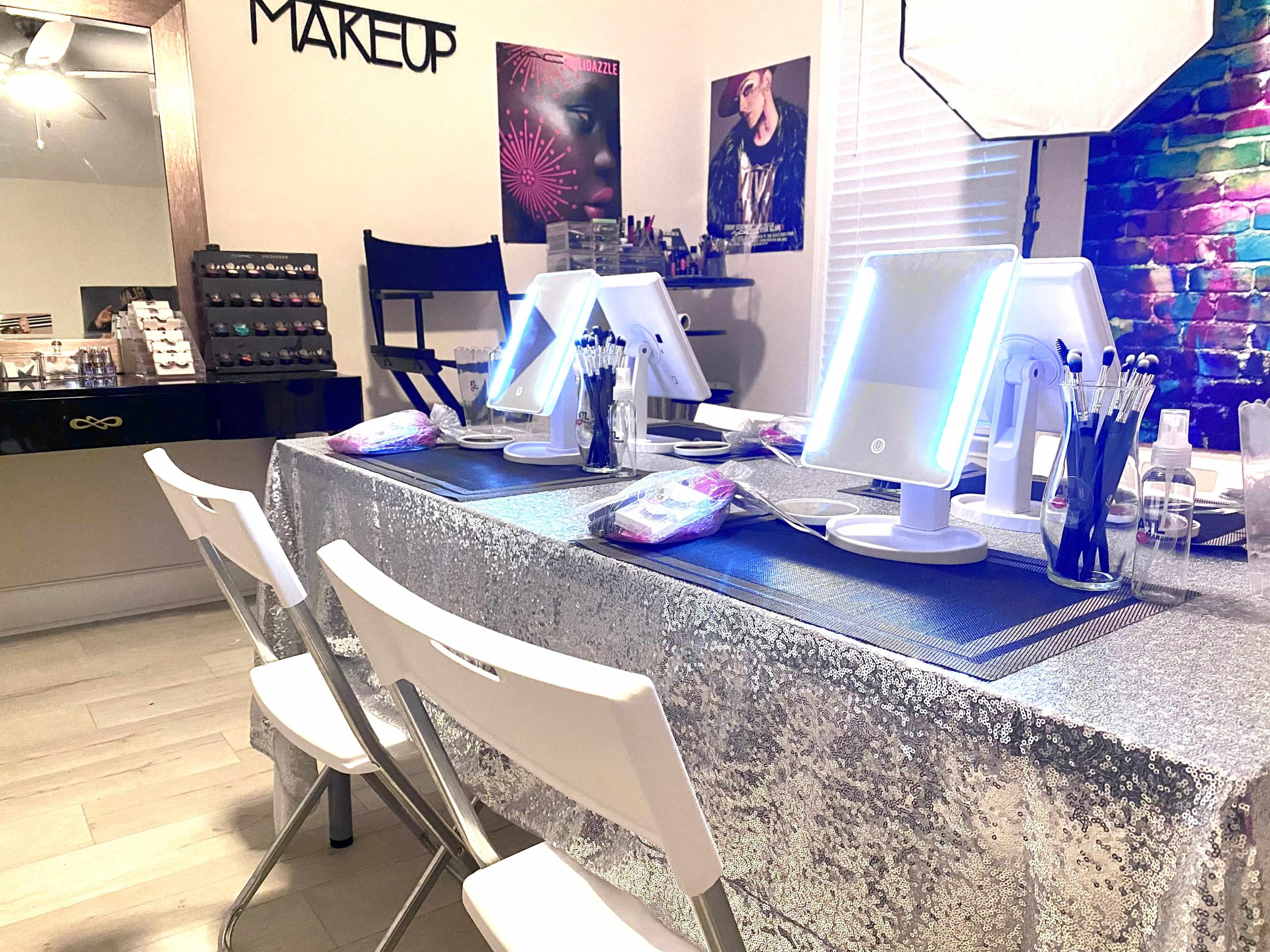 The image shows a makeup studio with a glittering table set up for clients, featuring illuminated mirrors and organized makeup tools, alongside chairs and a display of makeup products on the wall.