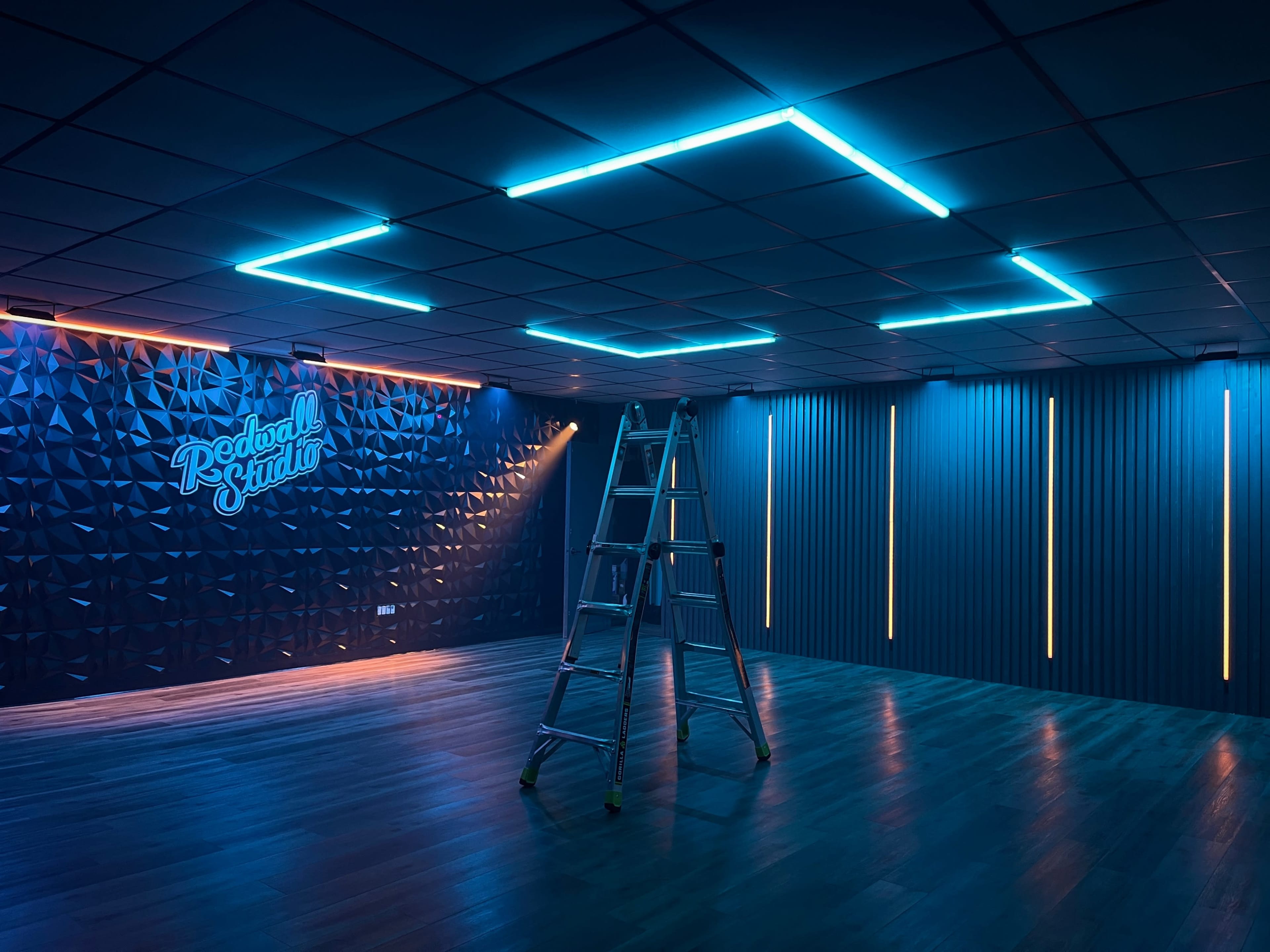 A brightly lit studio space features blue neon ceiling lights, a ladder, and textured wall panels with branding.