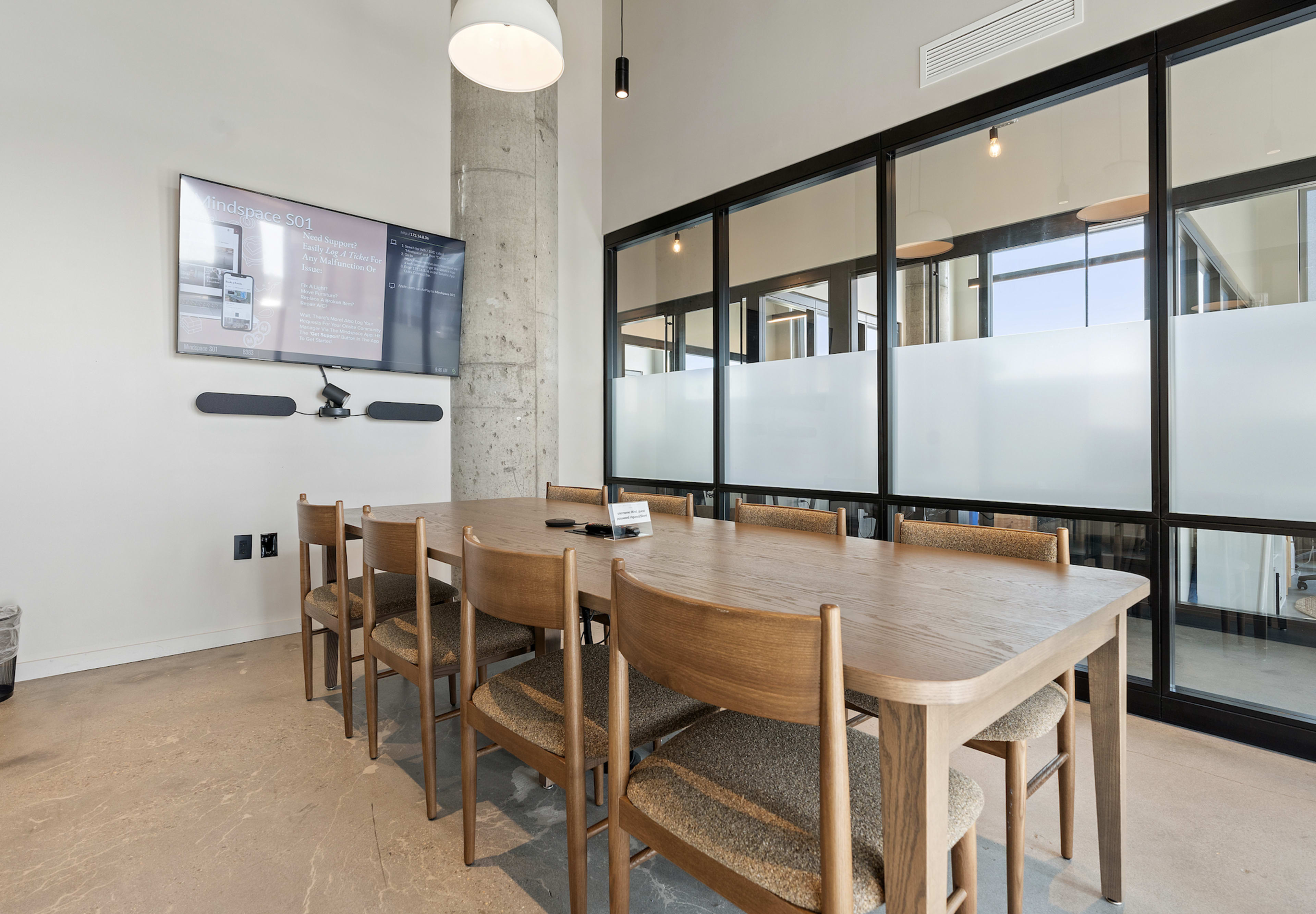 A conference room features a large wooden table surrounded by chairs, with a wall-mounted screen displaying information and large glass partitions.