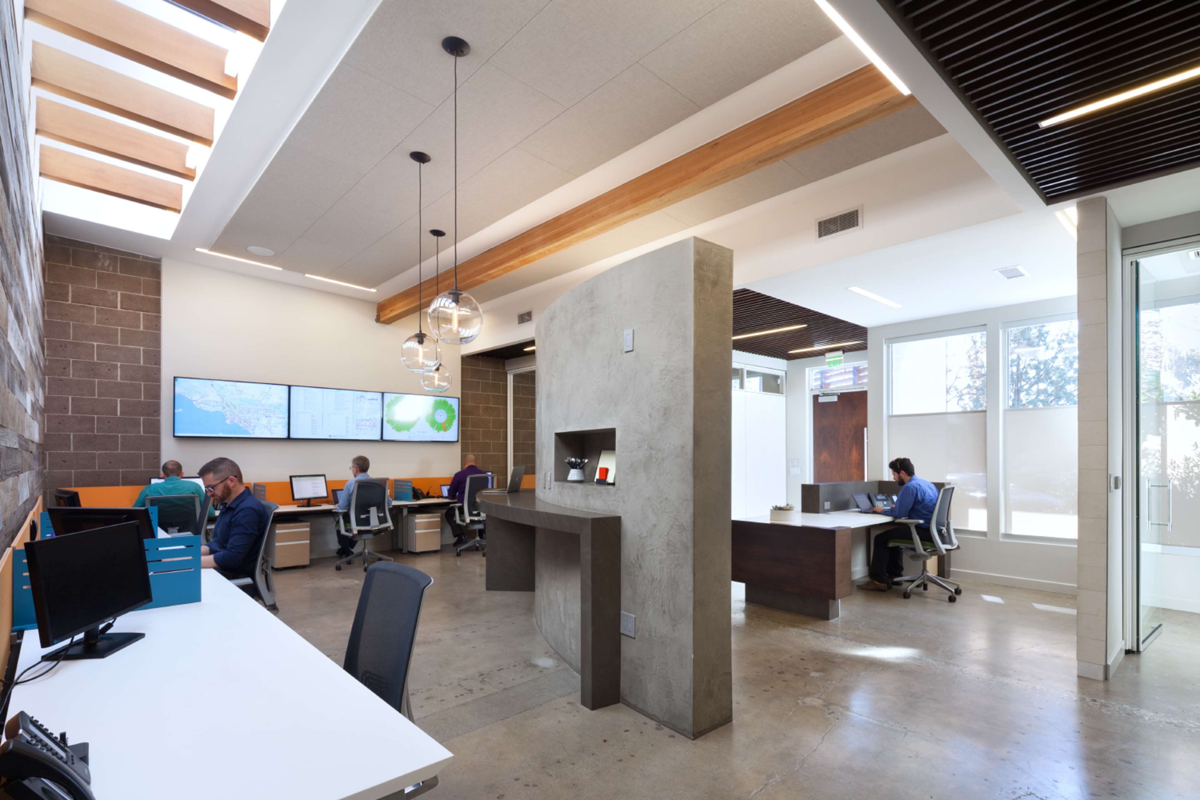 A modern open office space with several workstations, large screens displaying data, and a partially partitioned area featuring a central wall structure.