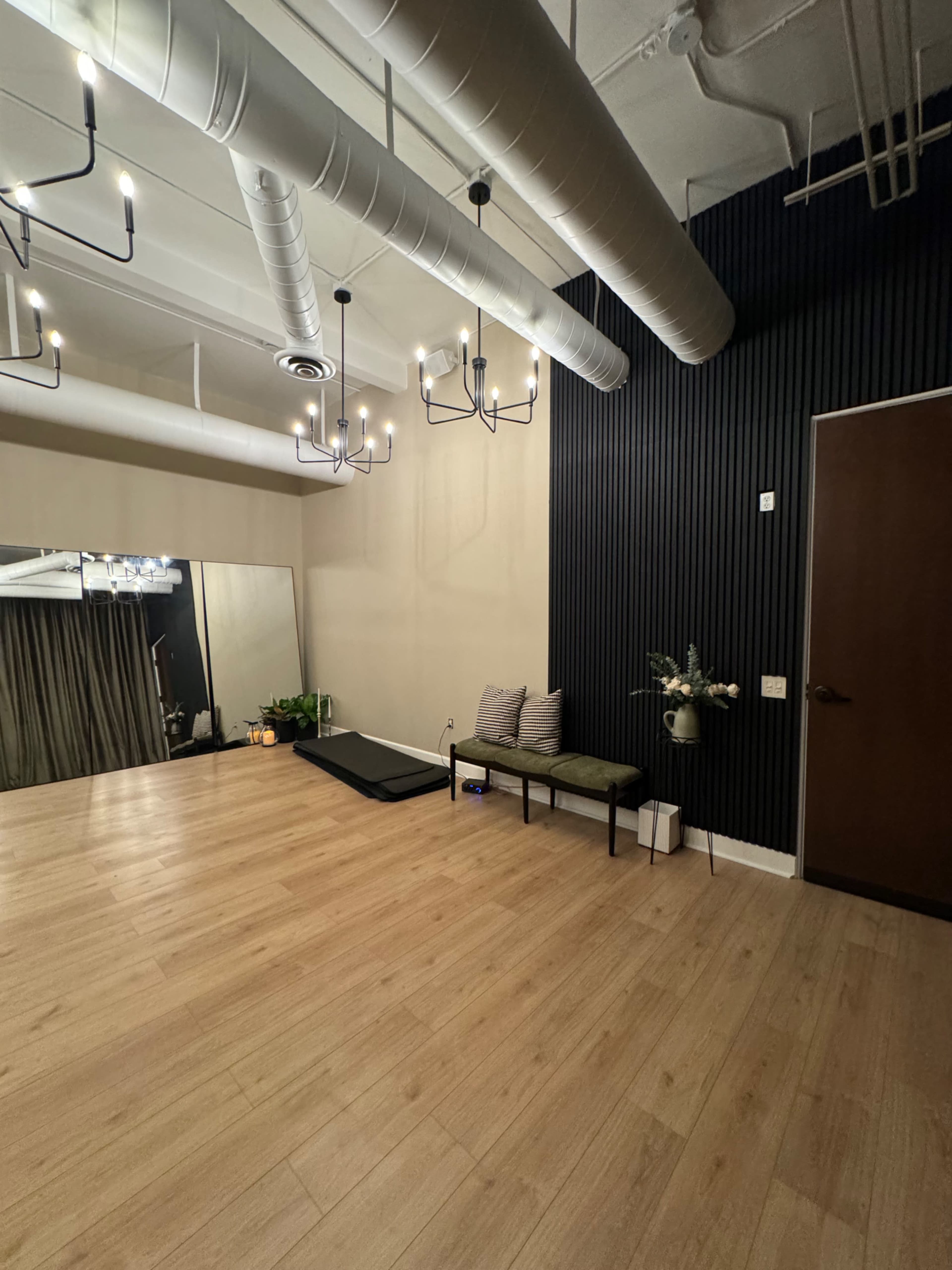 The image shows a spacious, modern room featuring wooden flooring, a dark accent wall, a bench with cushions, and a large mirror reflecting a curtained area.