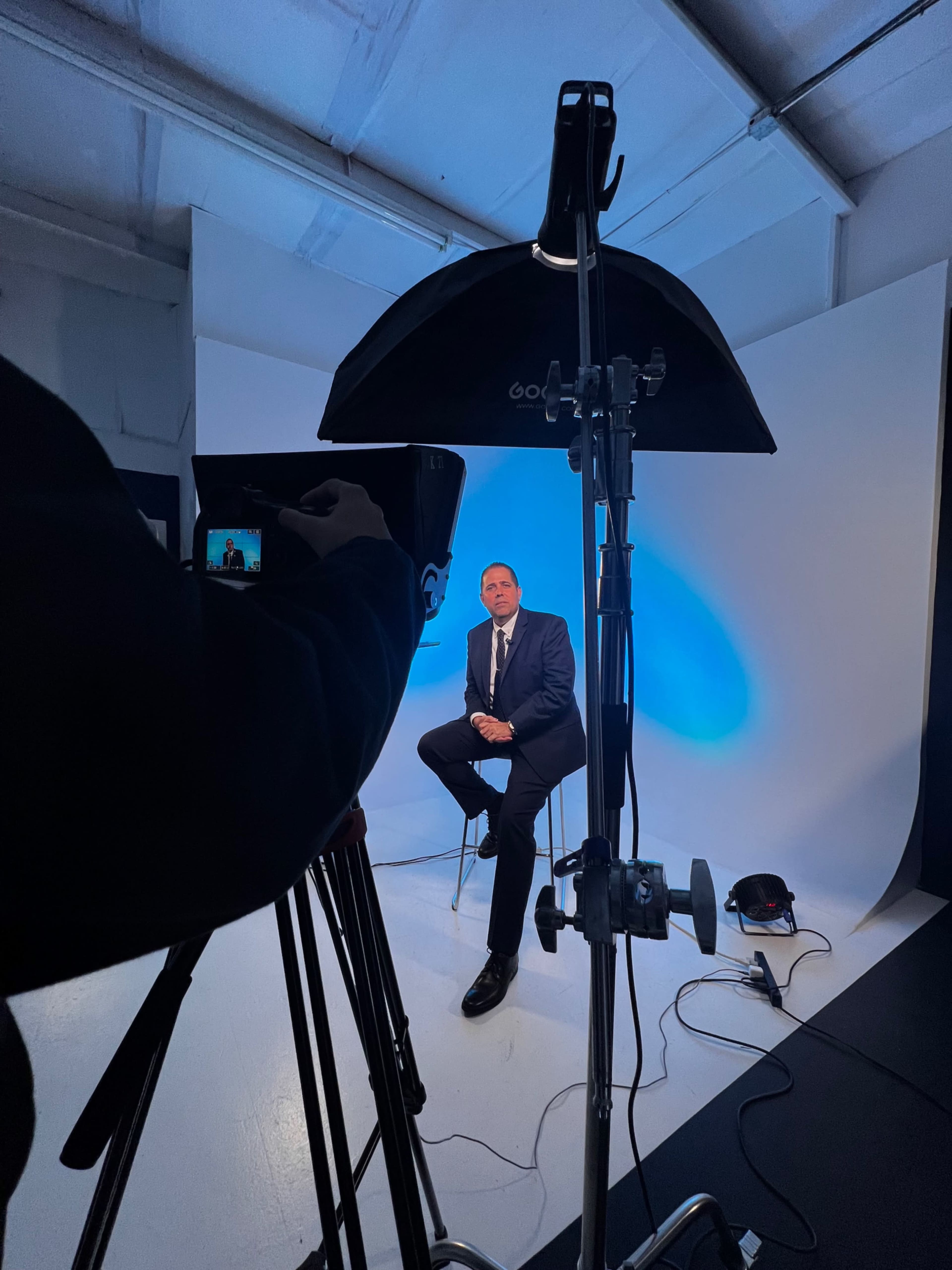 A man in a suit sits on a stool in front of a lit backdrop while a camera is positioned to capture the scene.