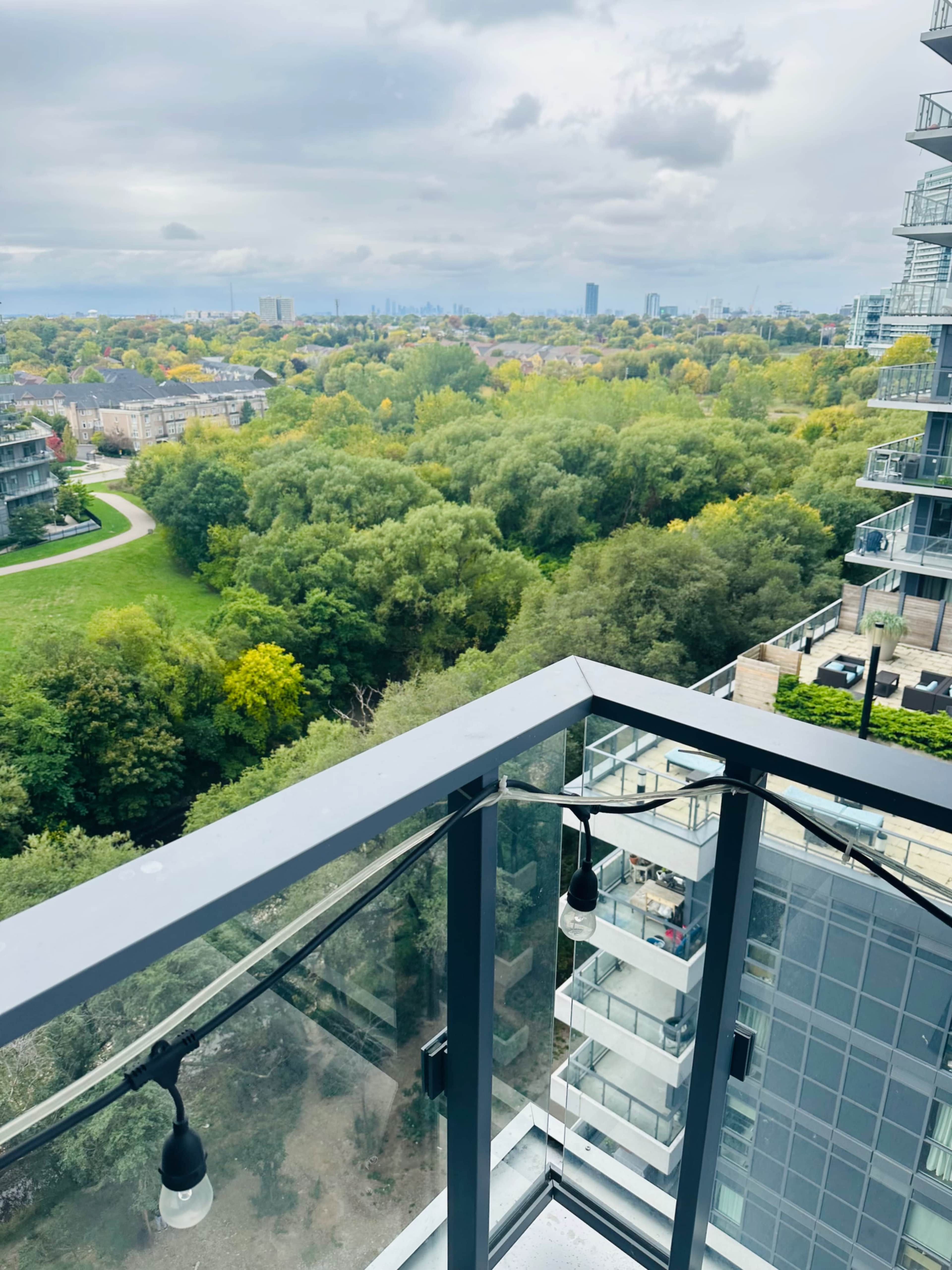 A balcony view overlooks a lush green landscape with trees, leading to a city skyline in the distance under a cloudy sky.