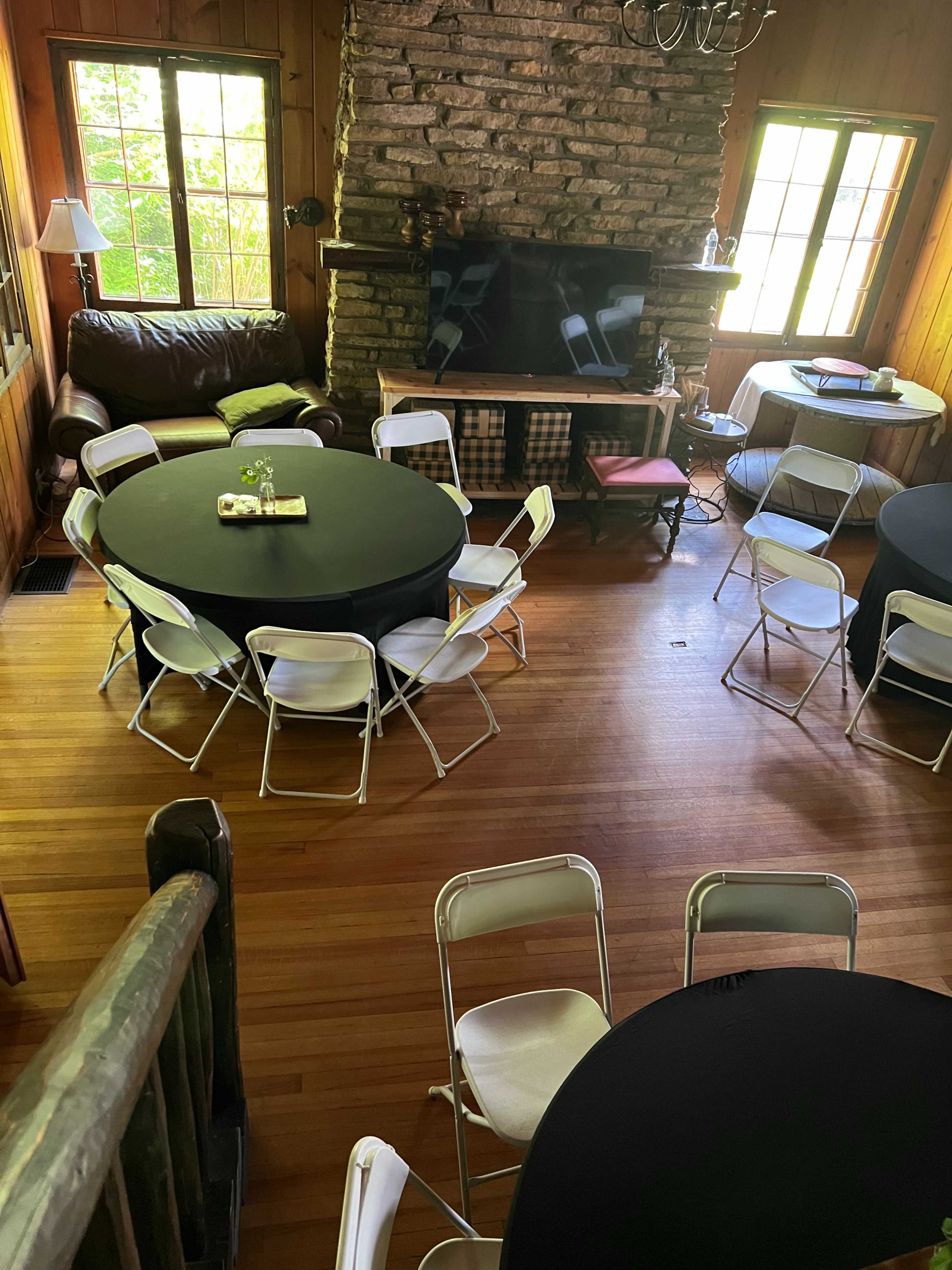 The image shows a spacious, wooden-paneled room arranged with black-covered tables and white folding chairs, featuring a stone fireplace and a television.