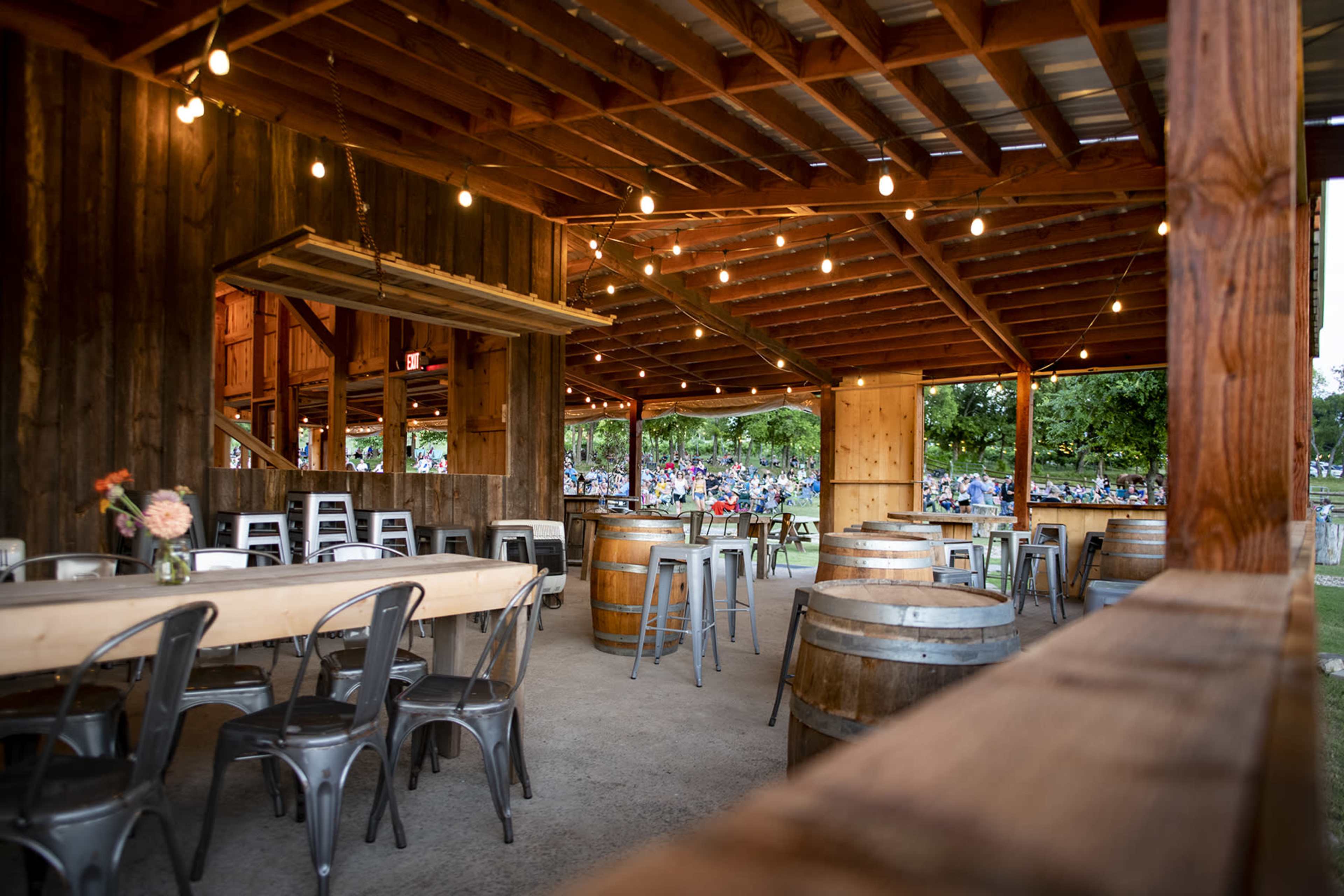 The image features a rustic interior of a barn-like venue with wooden beams, tables, and barrels, lit by string lights, creating a festive atmosphere.