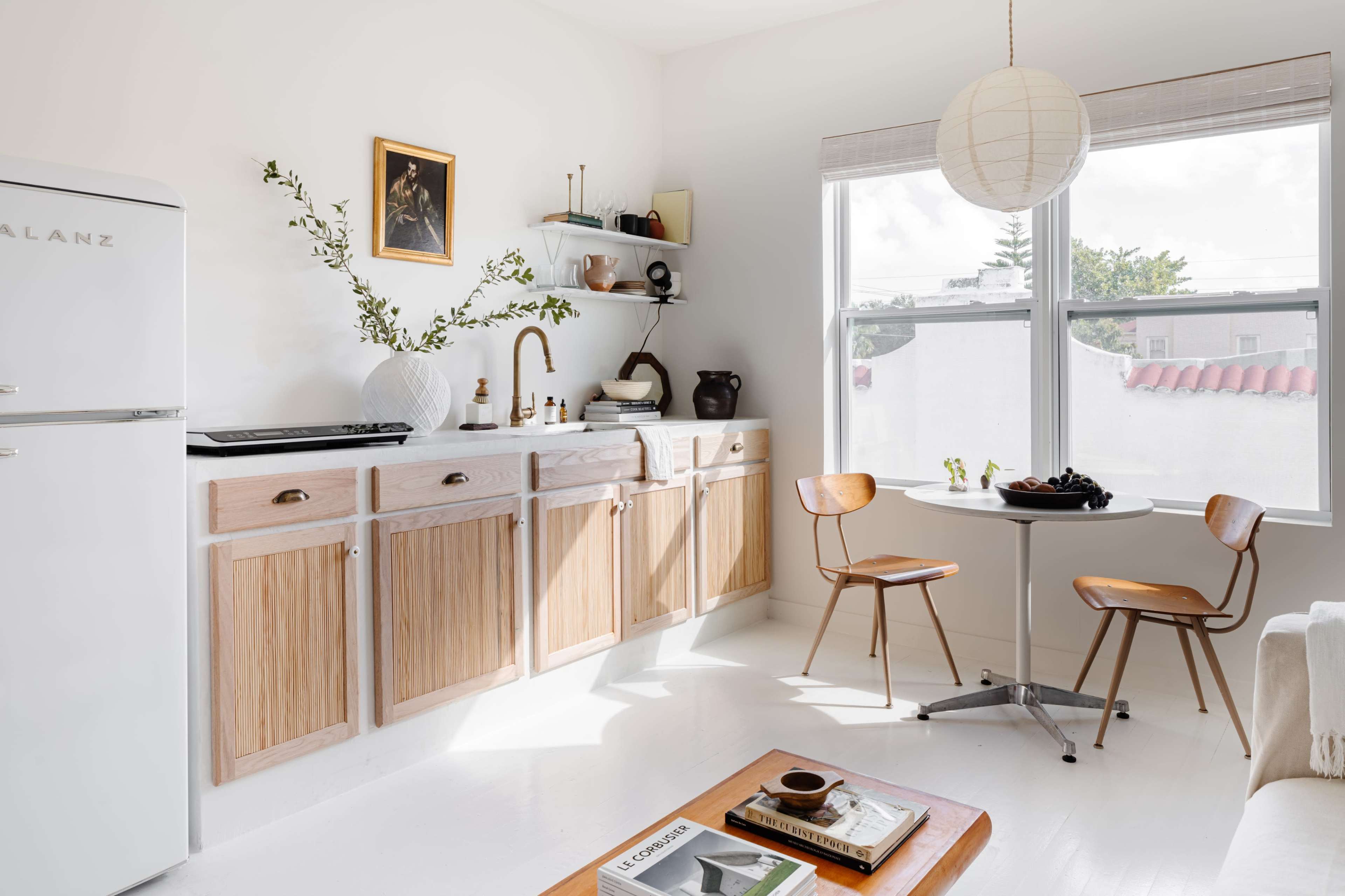 A minimalist kitchen features wooden cabinetry, a vintage refrigerator, and a small dining table with two chairs beside a window.