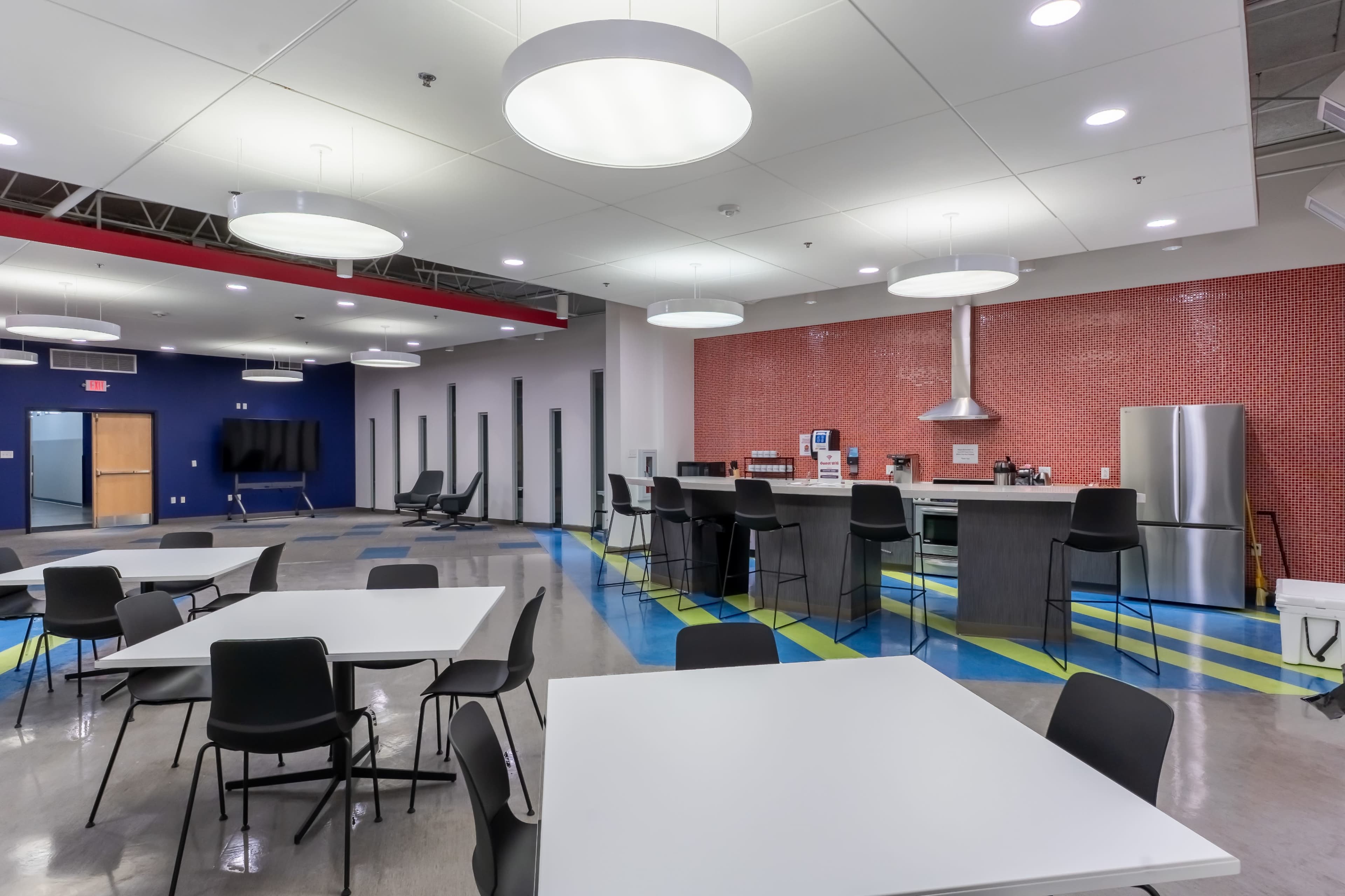 The image shows a modern communal kitchen and dining area with several tables and chairs, a kitchen counter, and a large refrigerator, set against a backdrop of colorful flooring and wall accents.