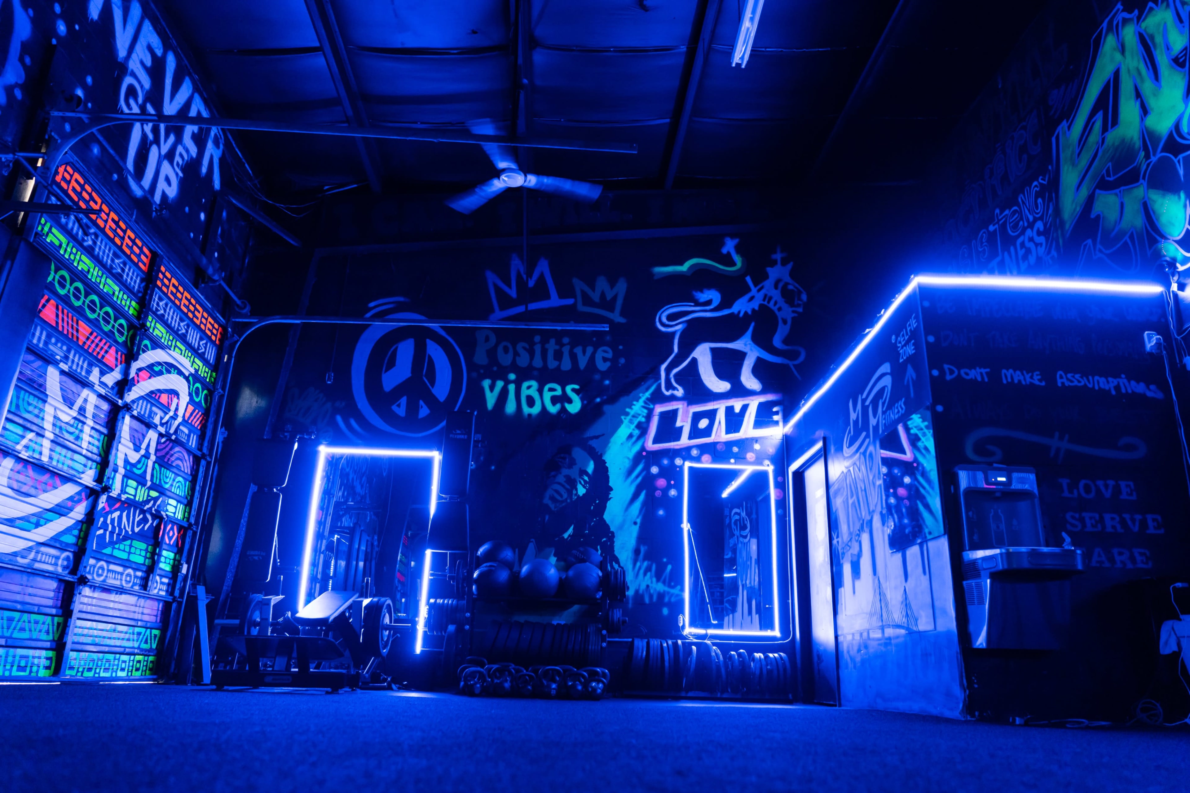 A vibrant gym interior with neon blue lighting, motivational murals on the walls, and exercise equipment visible on the floor.