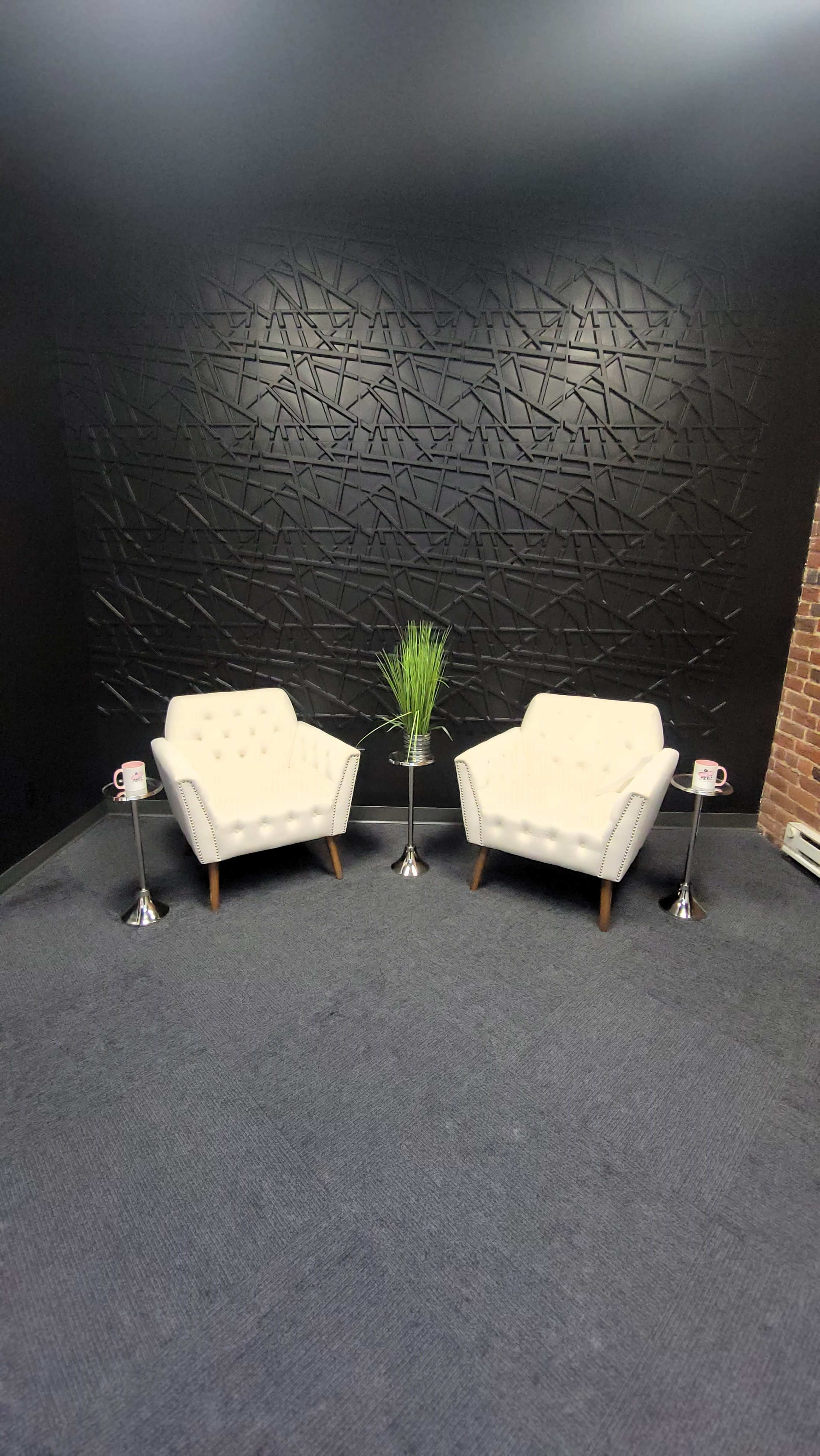 A small, modern seating area with two white chairs facing each other, a small table with decorative plants between them, and a textured black wall behind.
