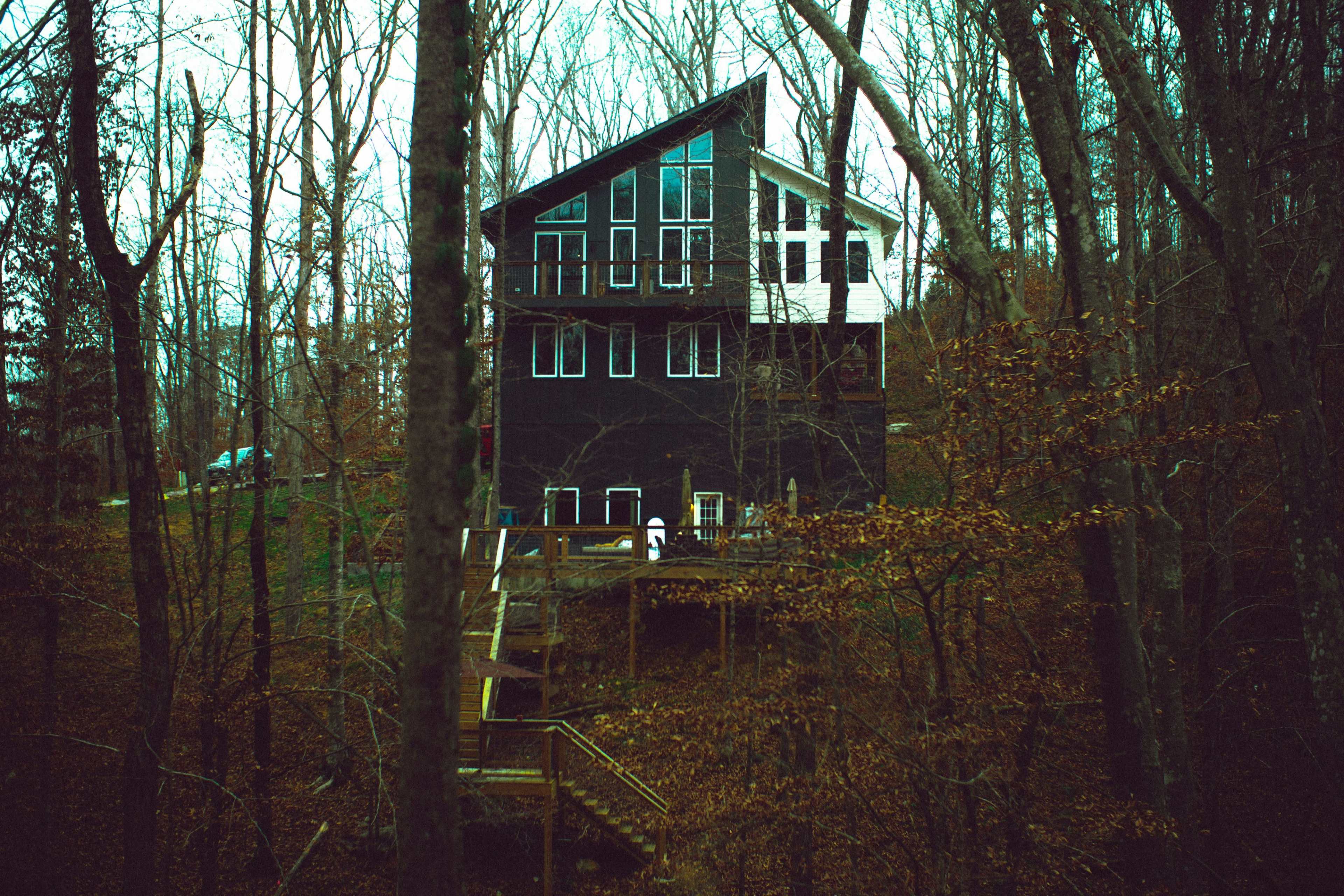 A tall, modern house with large windows is situated on a hillside, surrounded by bare trees in a wooded area.