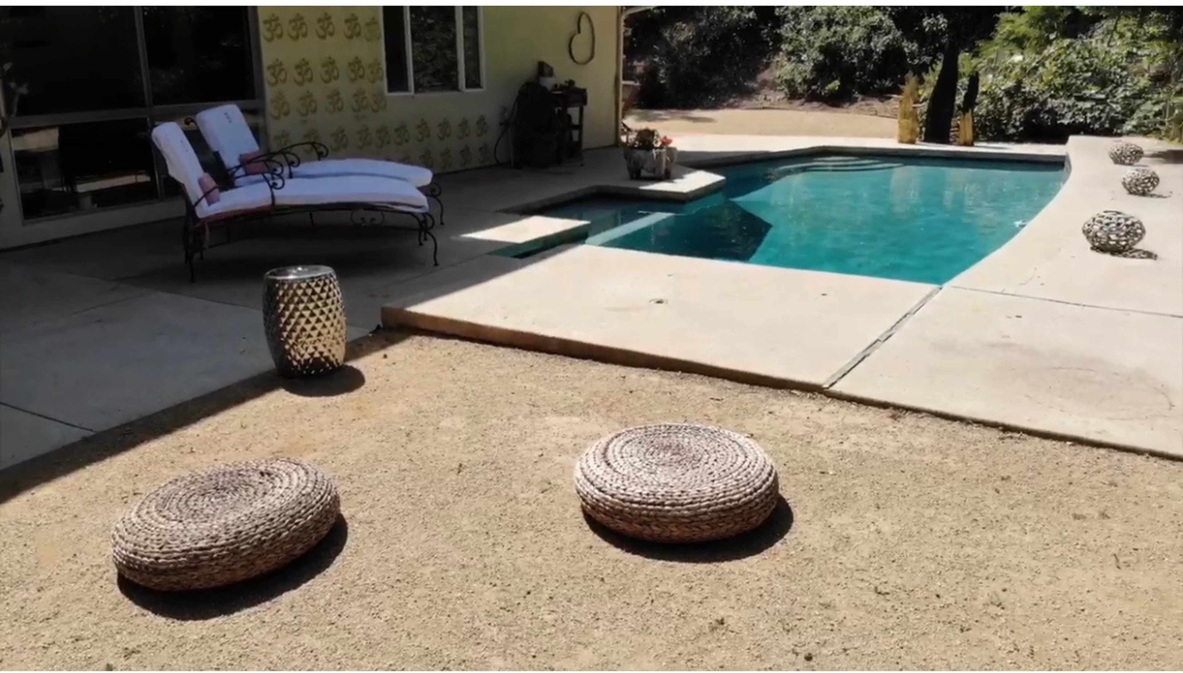 A pair of lounge chairs is positioned near a swimming pool, with woven round cushions placed on the ground nearby.