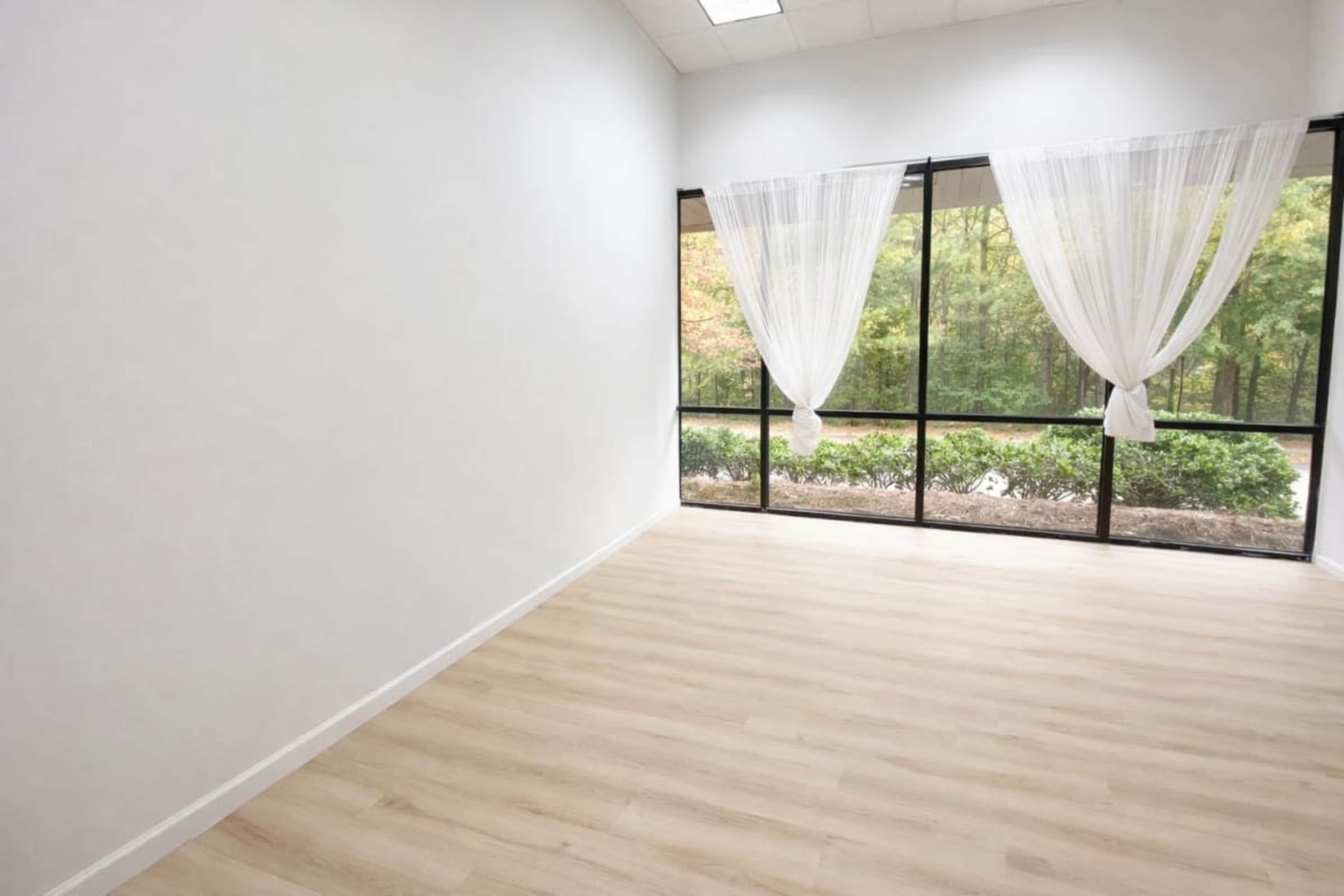 The image shows an empty room with light wood flooring and large windows adorned with sheer white curtains, overlooking a green landscape.