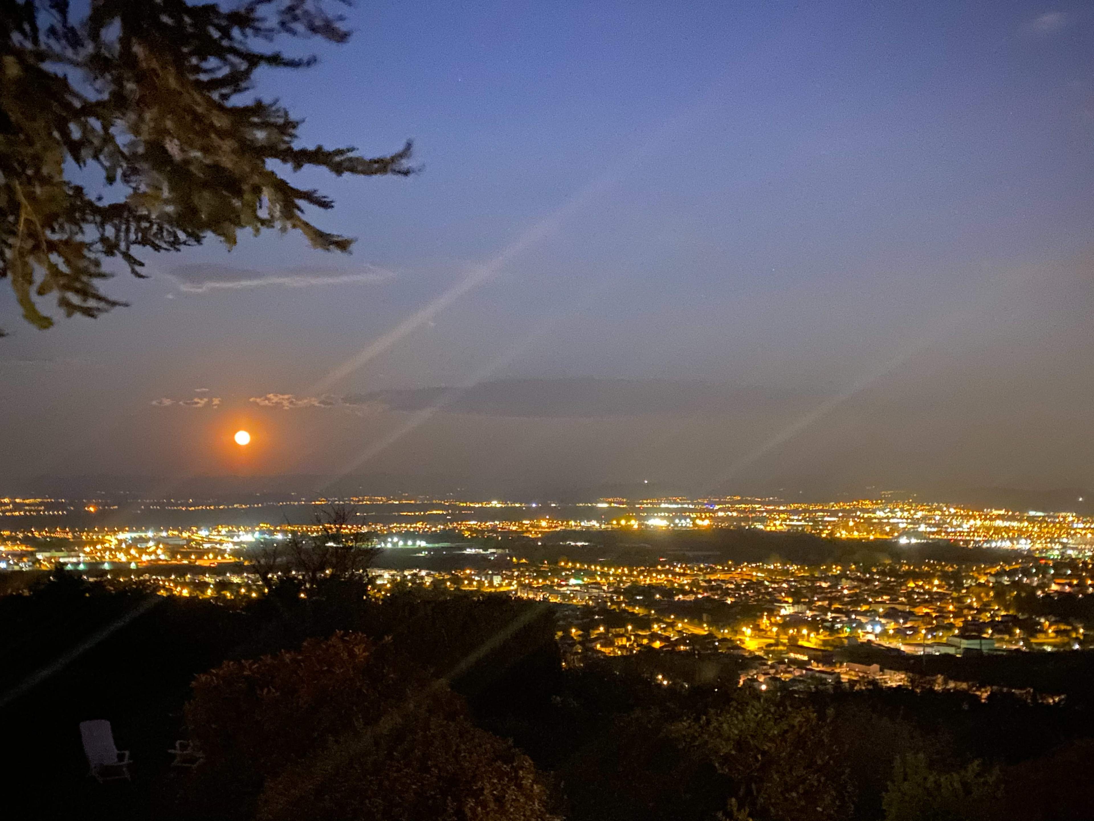 A view of a city illuminated at night, featuring a large, glowing moon rising above the horizon.
