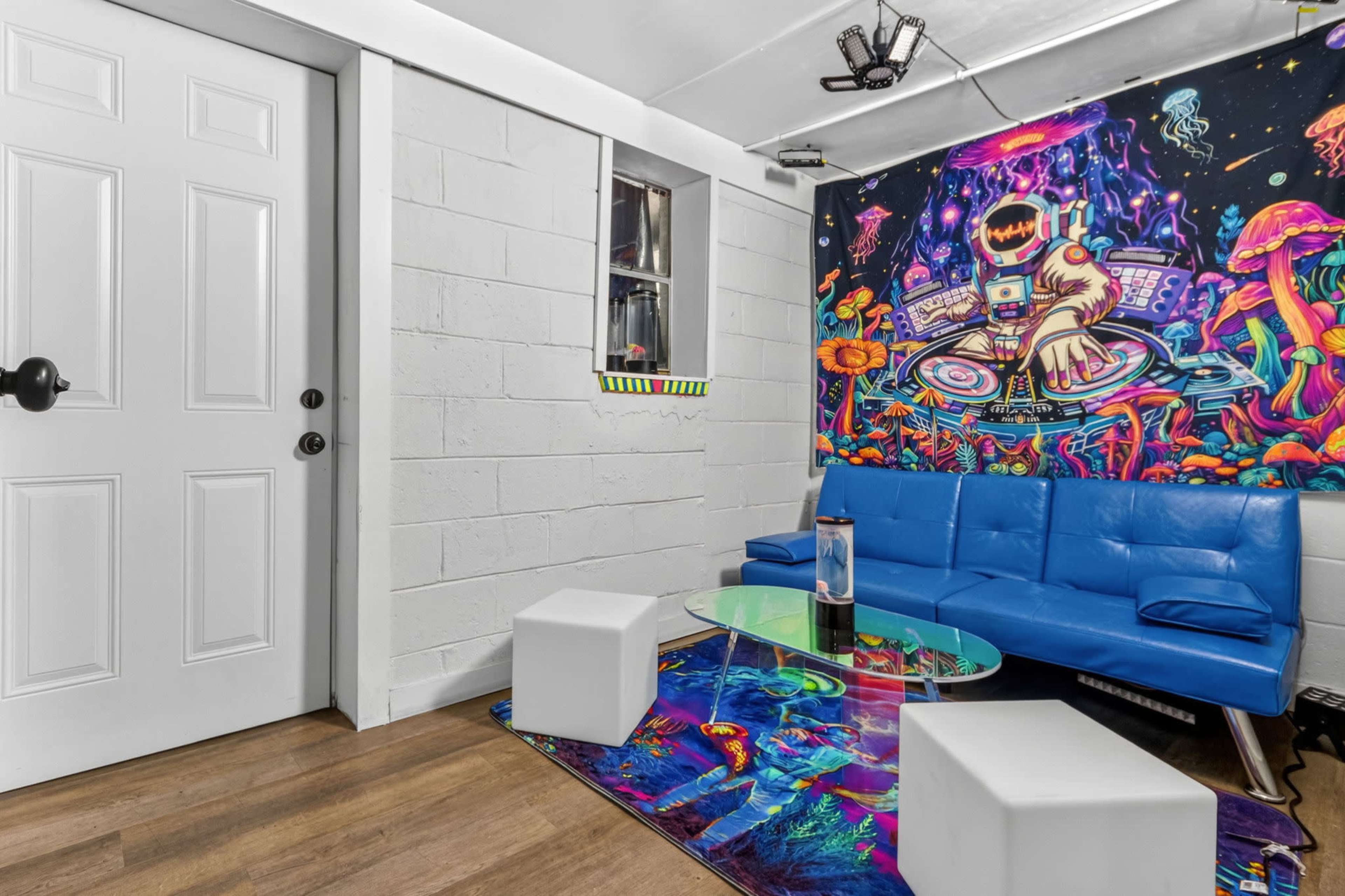 The image shows a brightly decorated room featuring a blue couch, a glass coffee table, and a vibrant mural of an astronaut surrounded by colorful mushrooms and abstract designs on one wall.