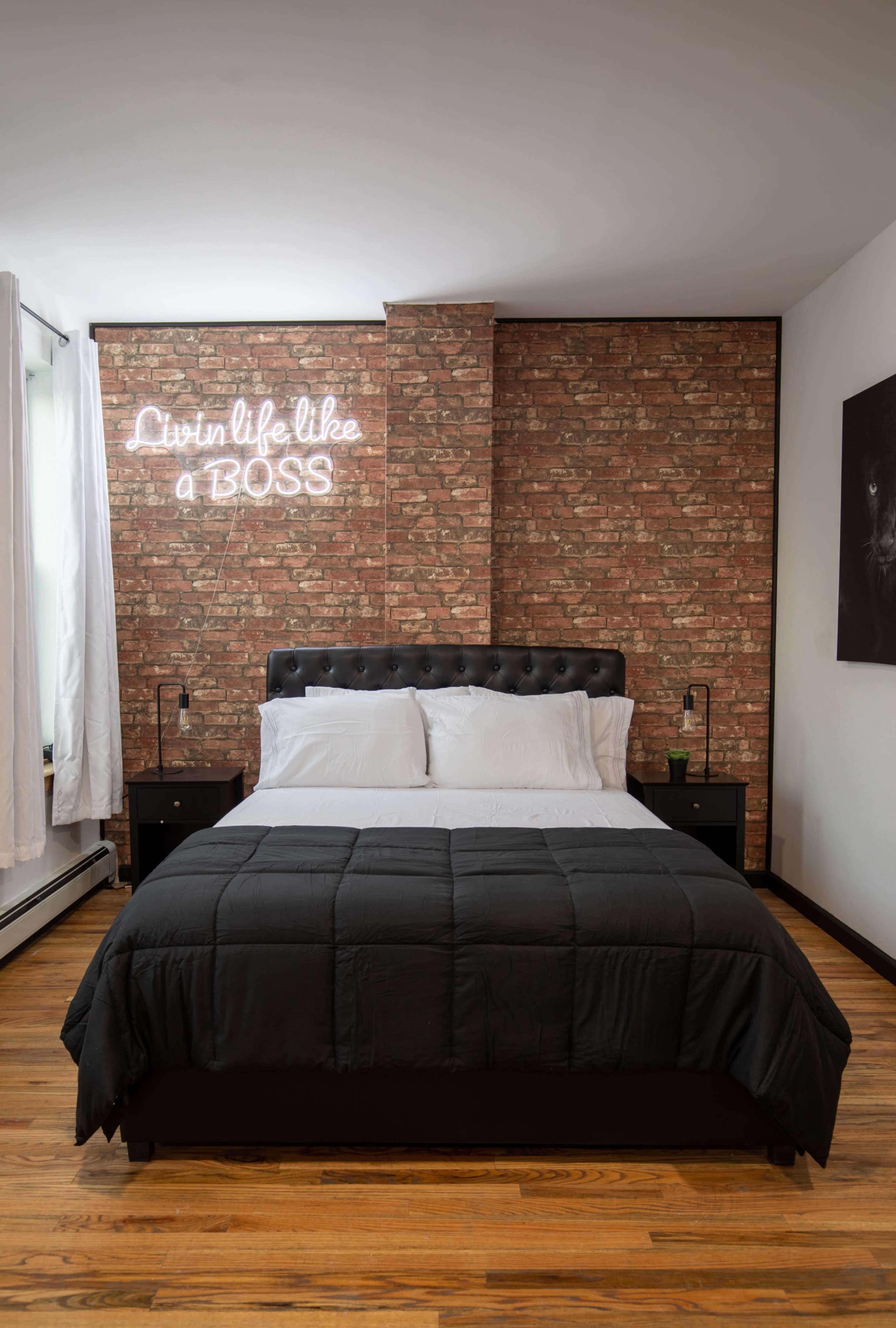A modern bedroom with a black bedspread, a brick accent wall, and a neon sign that reads "Livin' life like a BOSS."