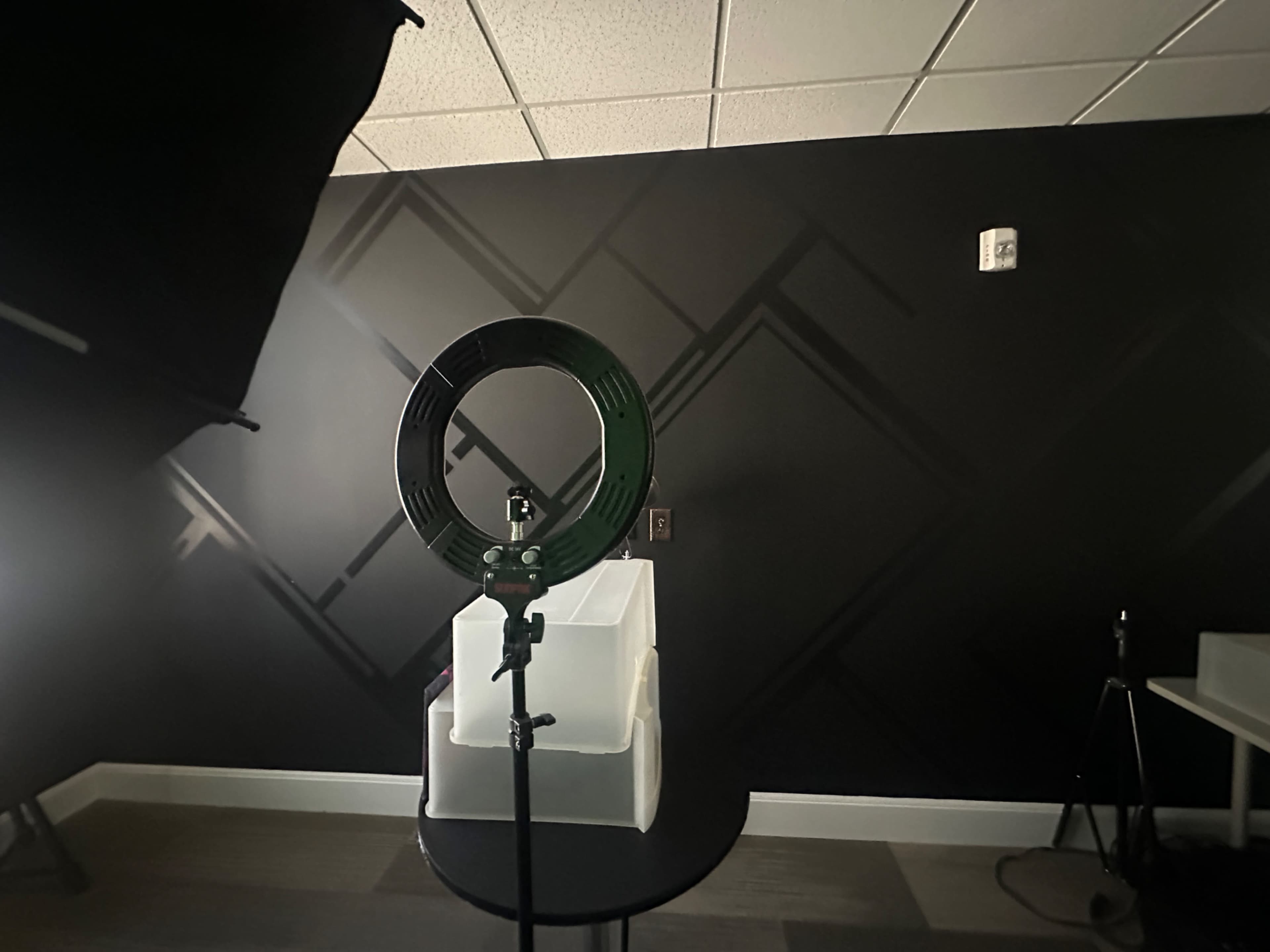 A ring light is positioned on a stand in front of a white structure on a round table, with a dark wall featuring a geometric design in the background.