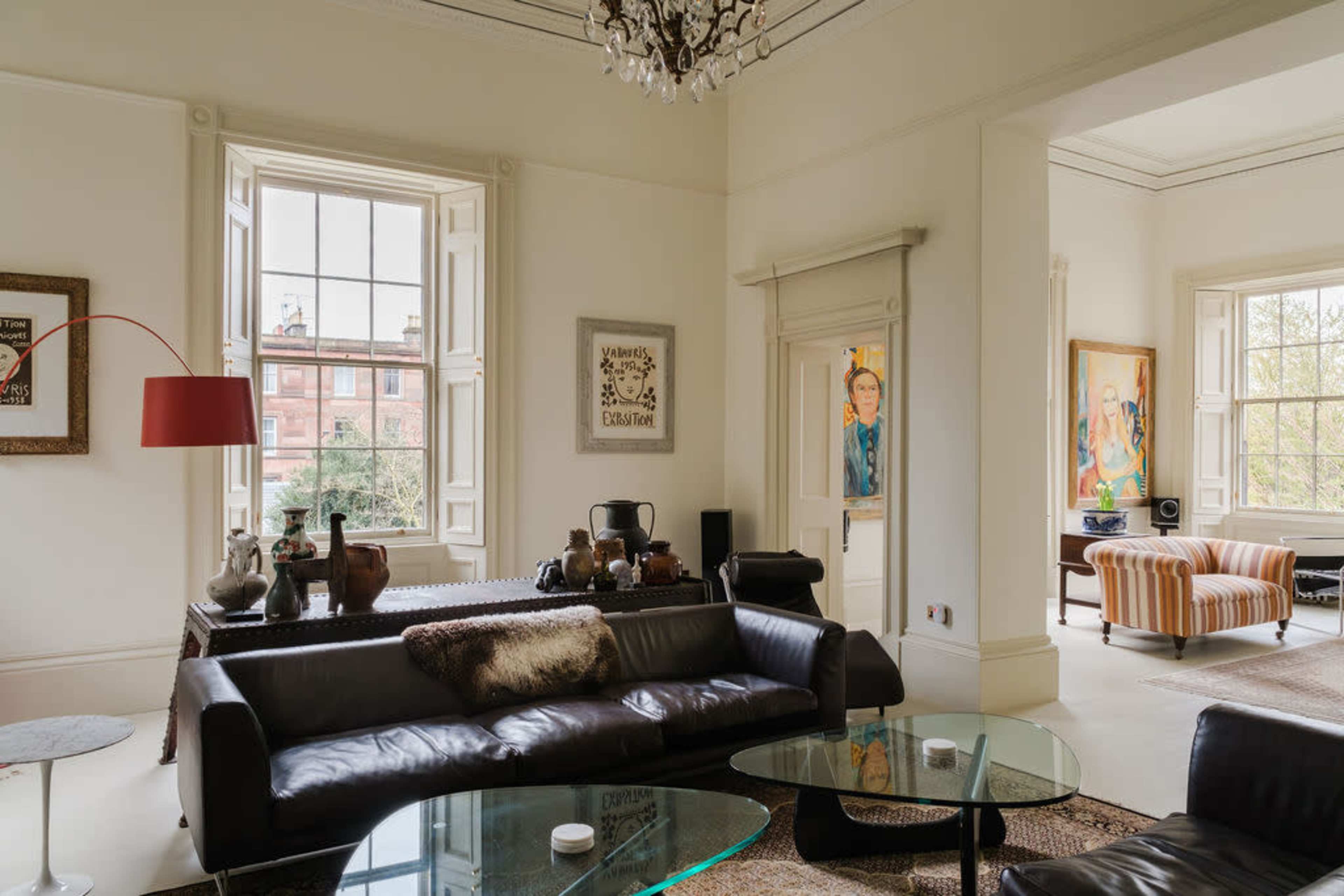 The image shows a spacious living room featuring a dark leather sofa, glass coffee tables, and large windows with natural light illuminating the space.
