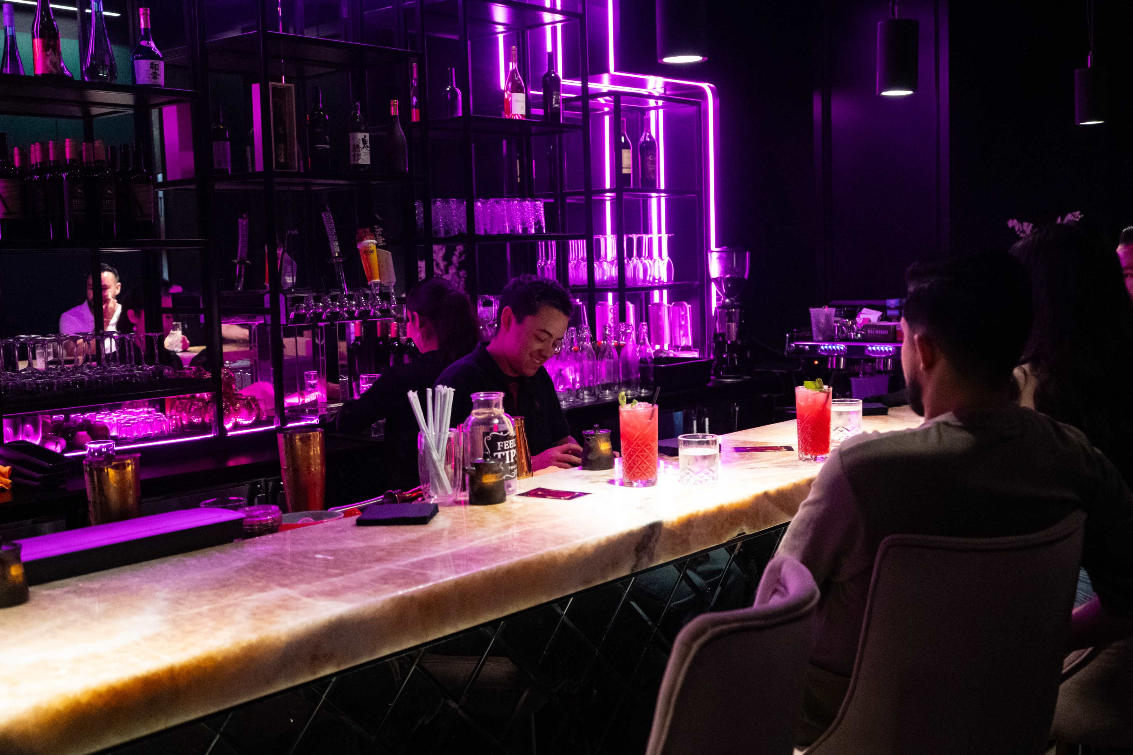 A bartender prepares drinks at a modern bar illuminated by purple lighting while customers sit at the counter.