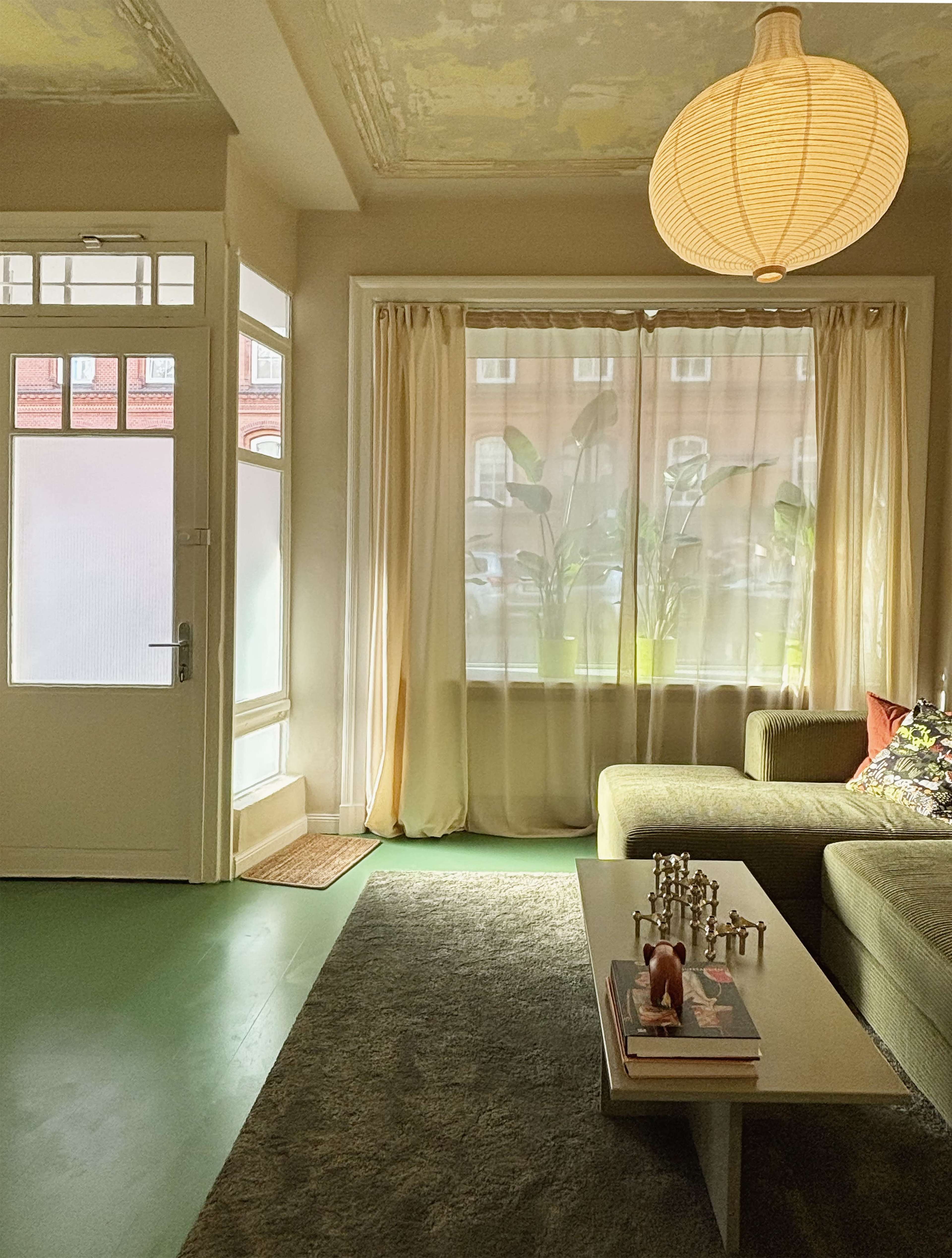 The image shows a cozy living room with a green sofa, a low coffee table adorned with decorative items, and large windows draped in light curtains letting in natural light.