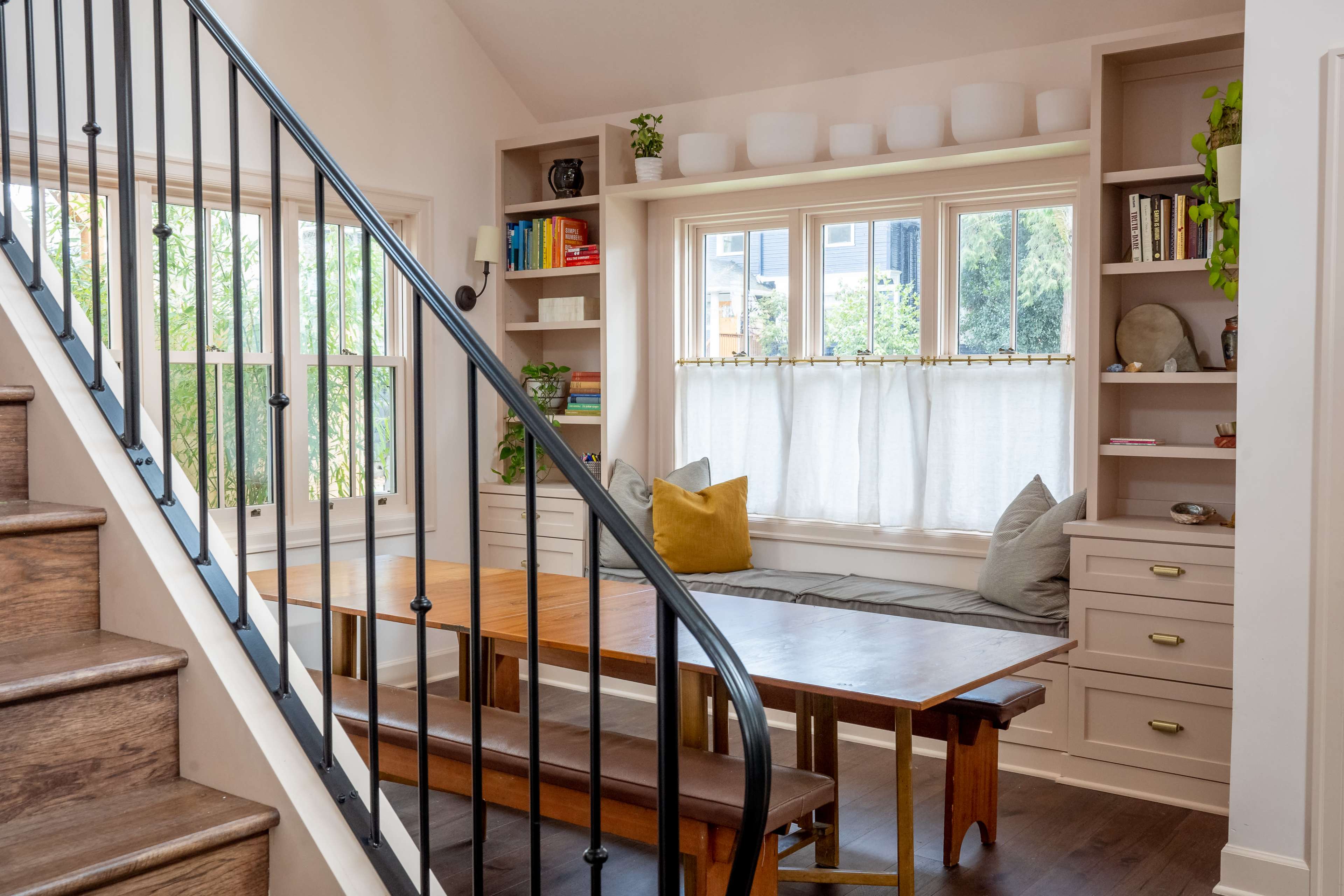 A well-lit interior space featuring a stairway, a dining table, and a cozy window seat surrounded by shelves and decorative items.