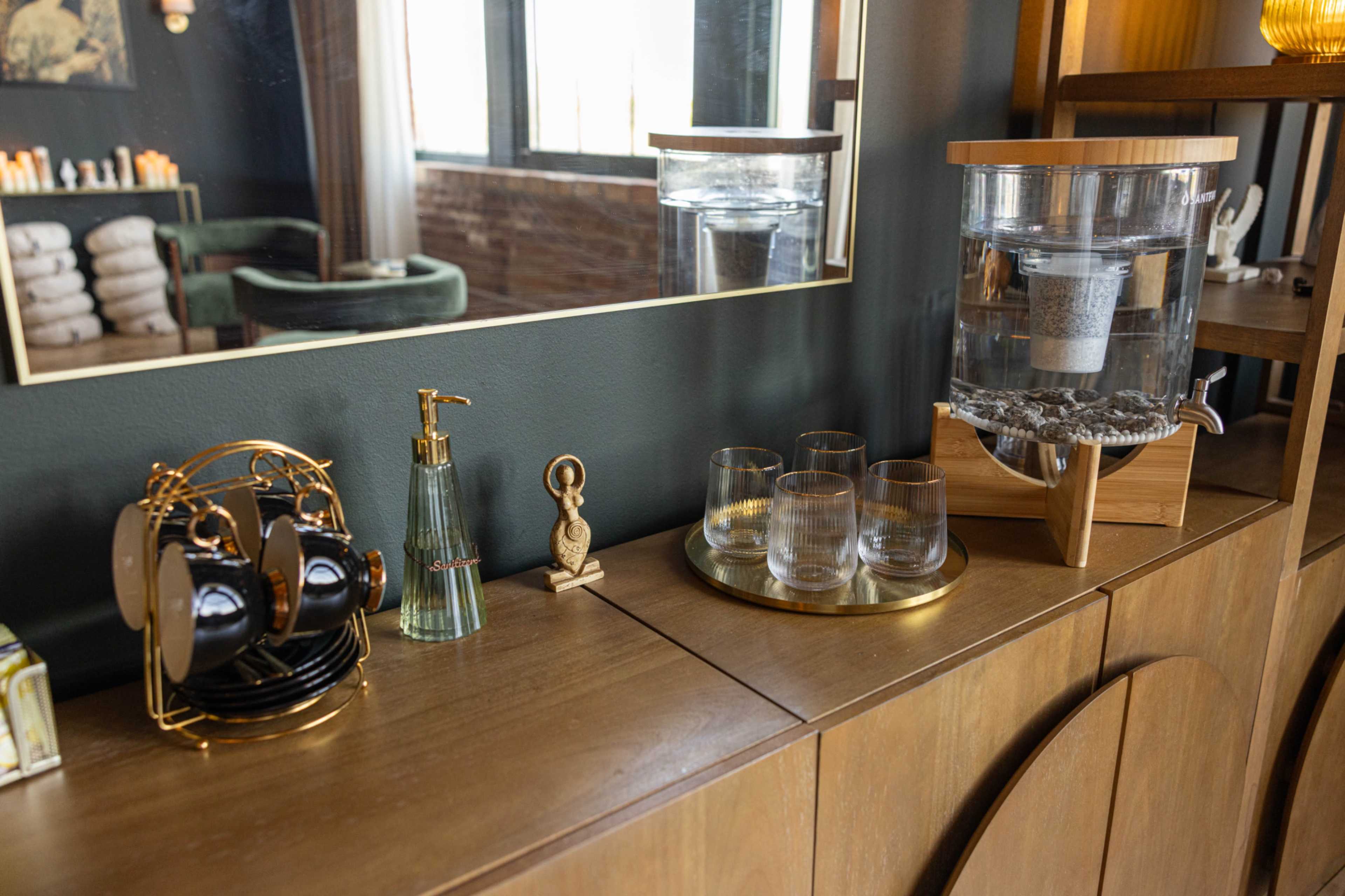 The image shows a wooden sideboard featuring a decorative water dispenser, glassware, and a holder for coffee cups, with a mirror reflecting the room's decor.
