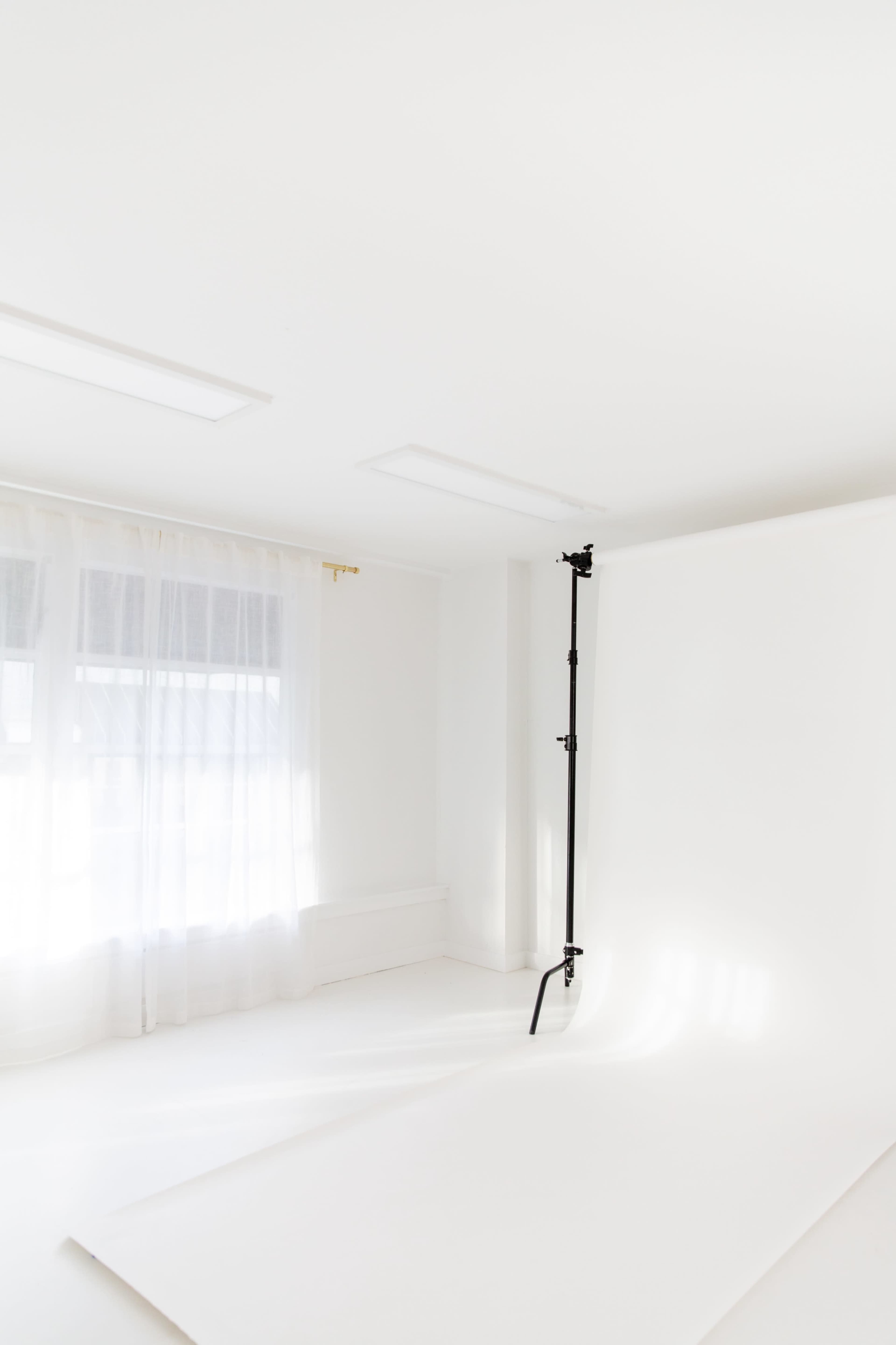 A bright, empty studio space features a large window with sheer curtains and a single black backdrop stand.