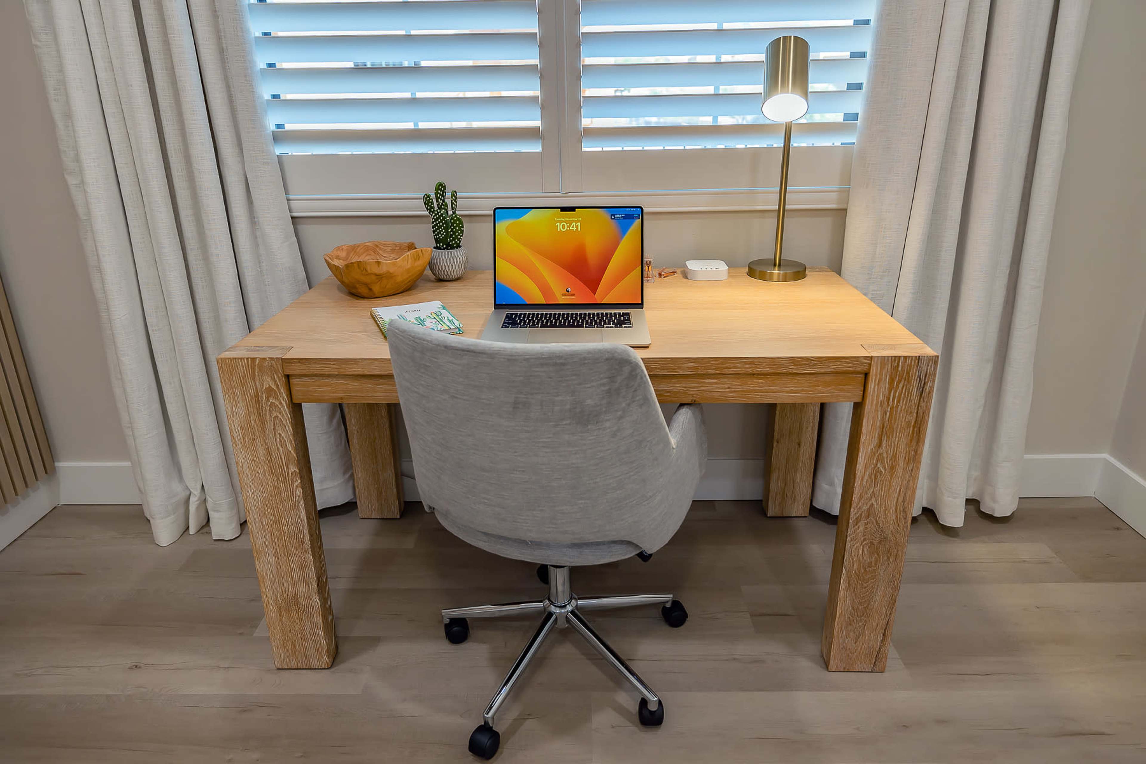 A wooden desk with a laptop, a lamp, a plant, and a bowl is positioned in front of a window with shuttered blinds.