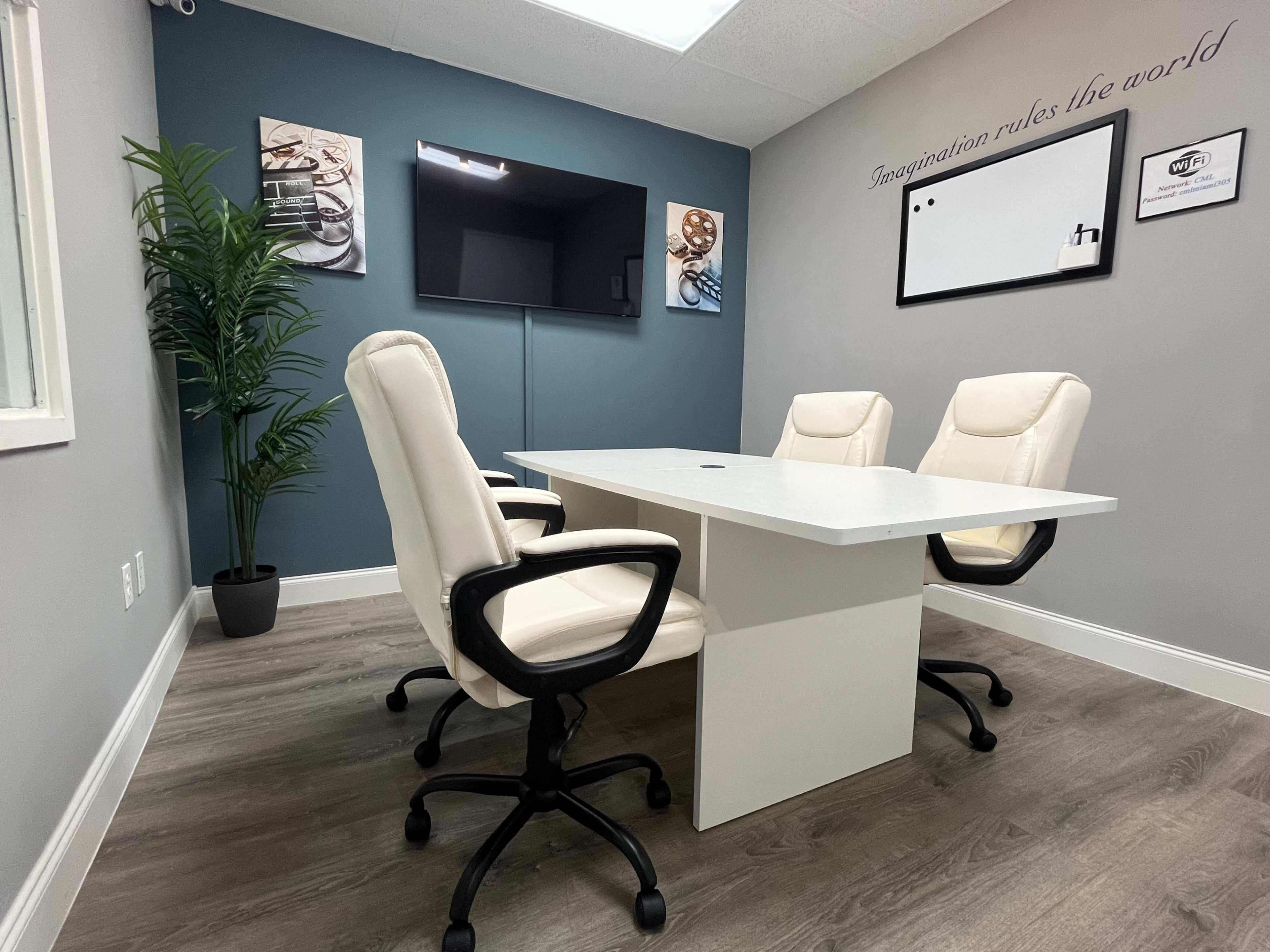 A modern conference room features a white table surrounded by three white ergonomic chairs, with a television mounted on the wall and a potted plant in the corner.