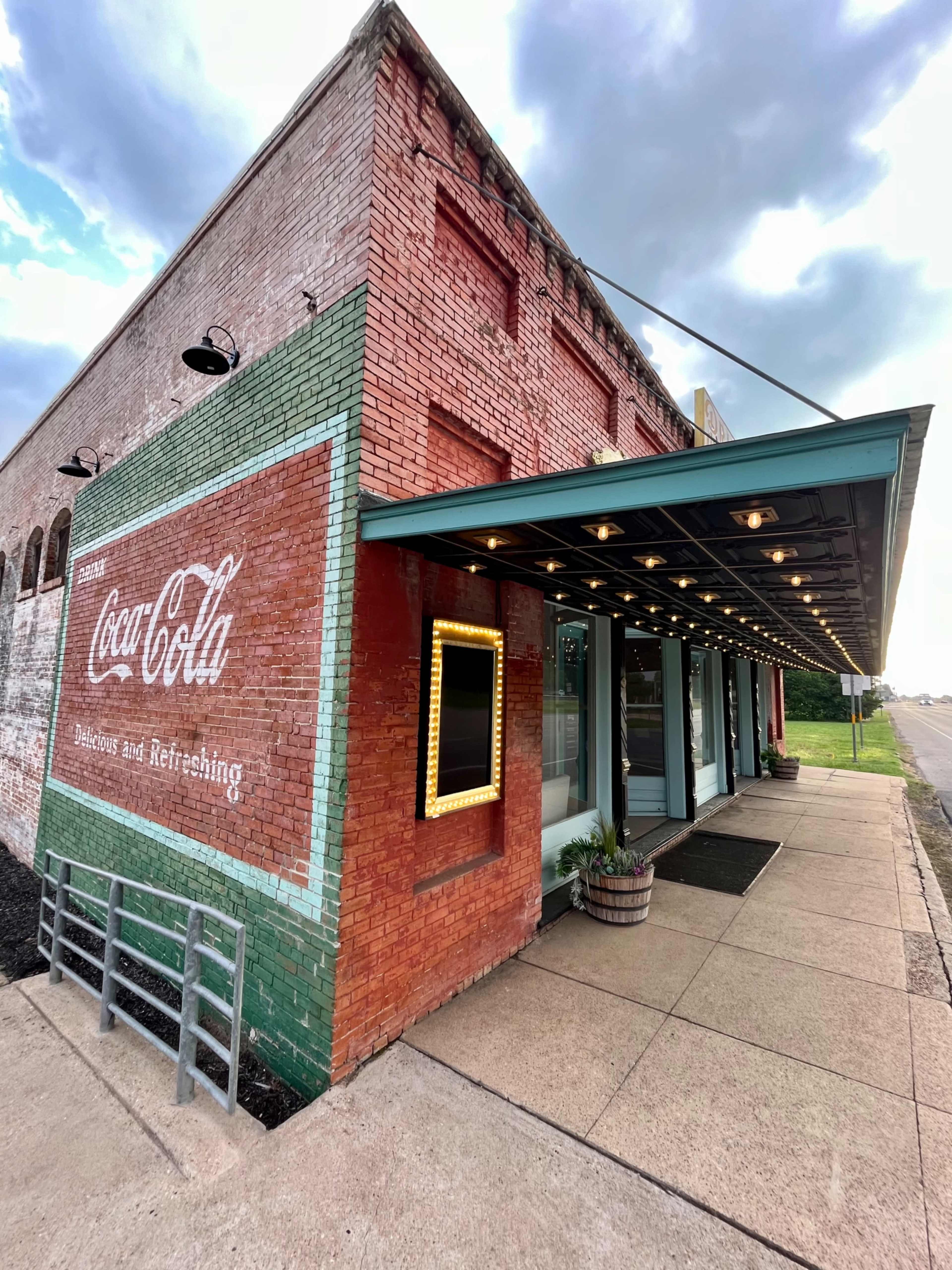 The image shows a brick building with a Coca-Cola advertisement on the side, featuring a covered entrance and a concrete walkway.